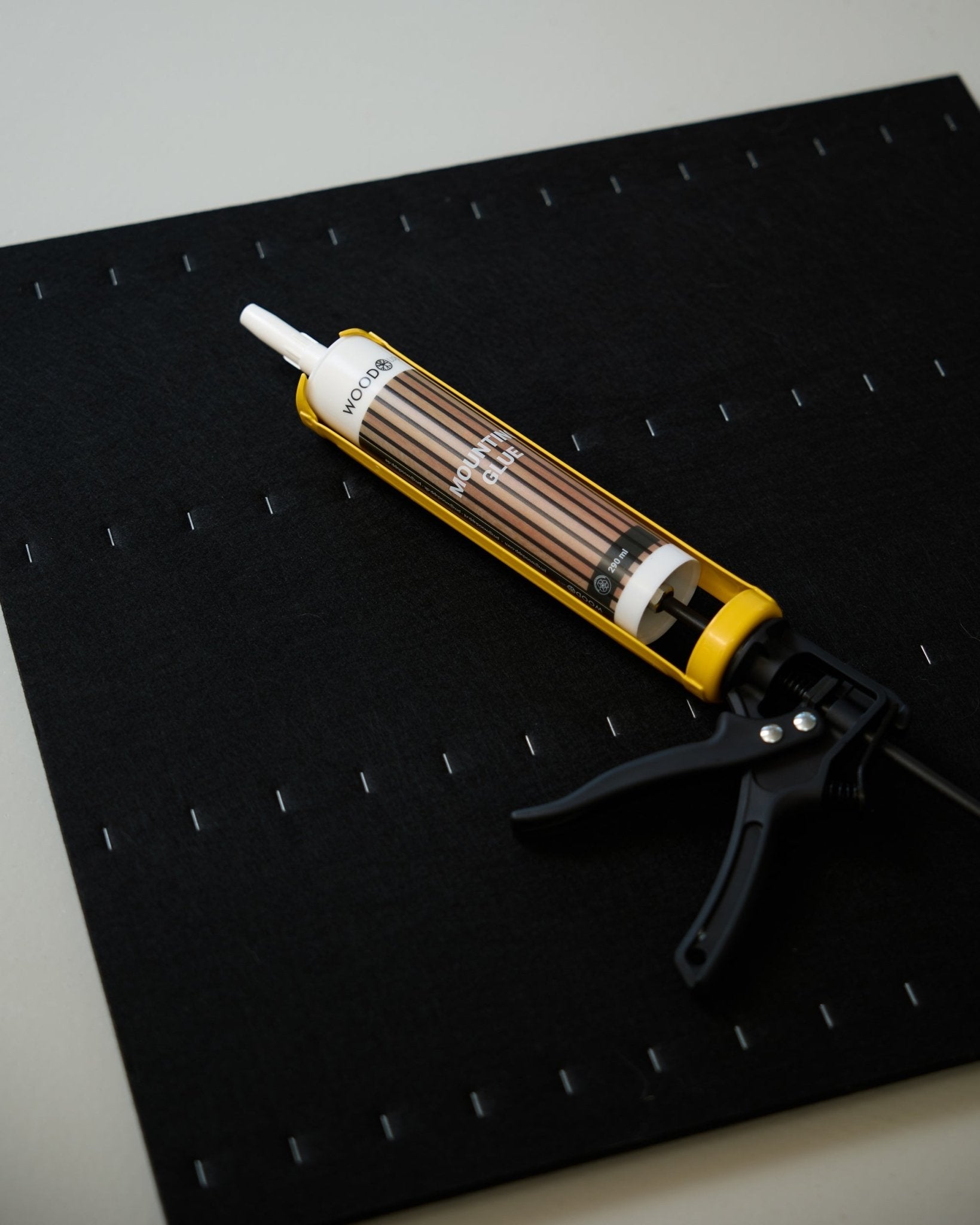 Un pistolet à mastic WoodUpp jaune et black (290 ml) repose sur un tapis black avec une grille d’agrafes blanches, utilisé pour installer des panneaux acoustiques.