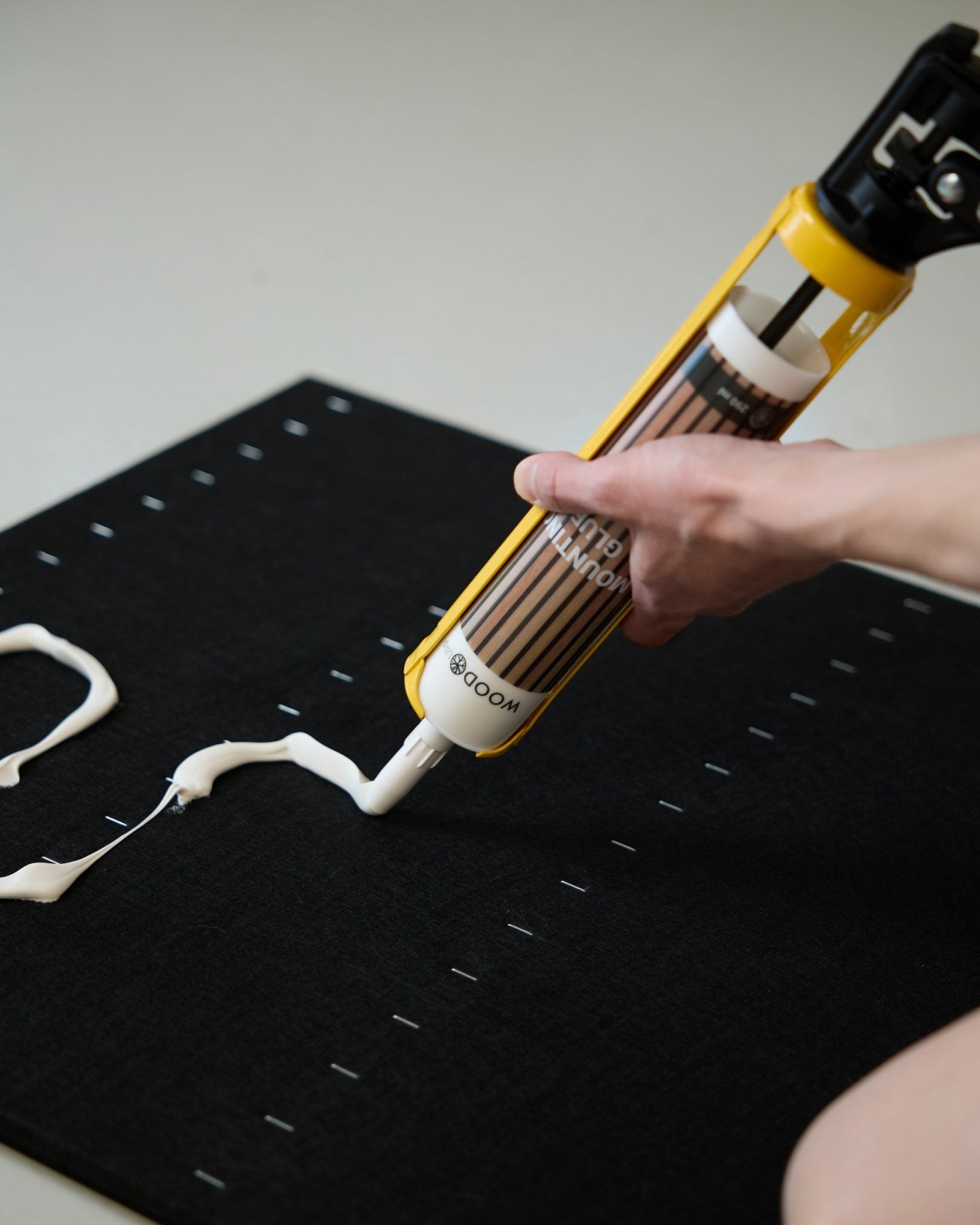 Une main applique de la colle de montage blanche en zigzag avec le pistolet à mastic WoodUpp (290 ml) sur une surface de panneau black Panneau acoustique.