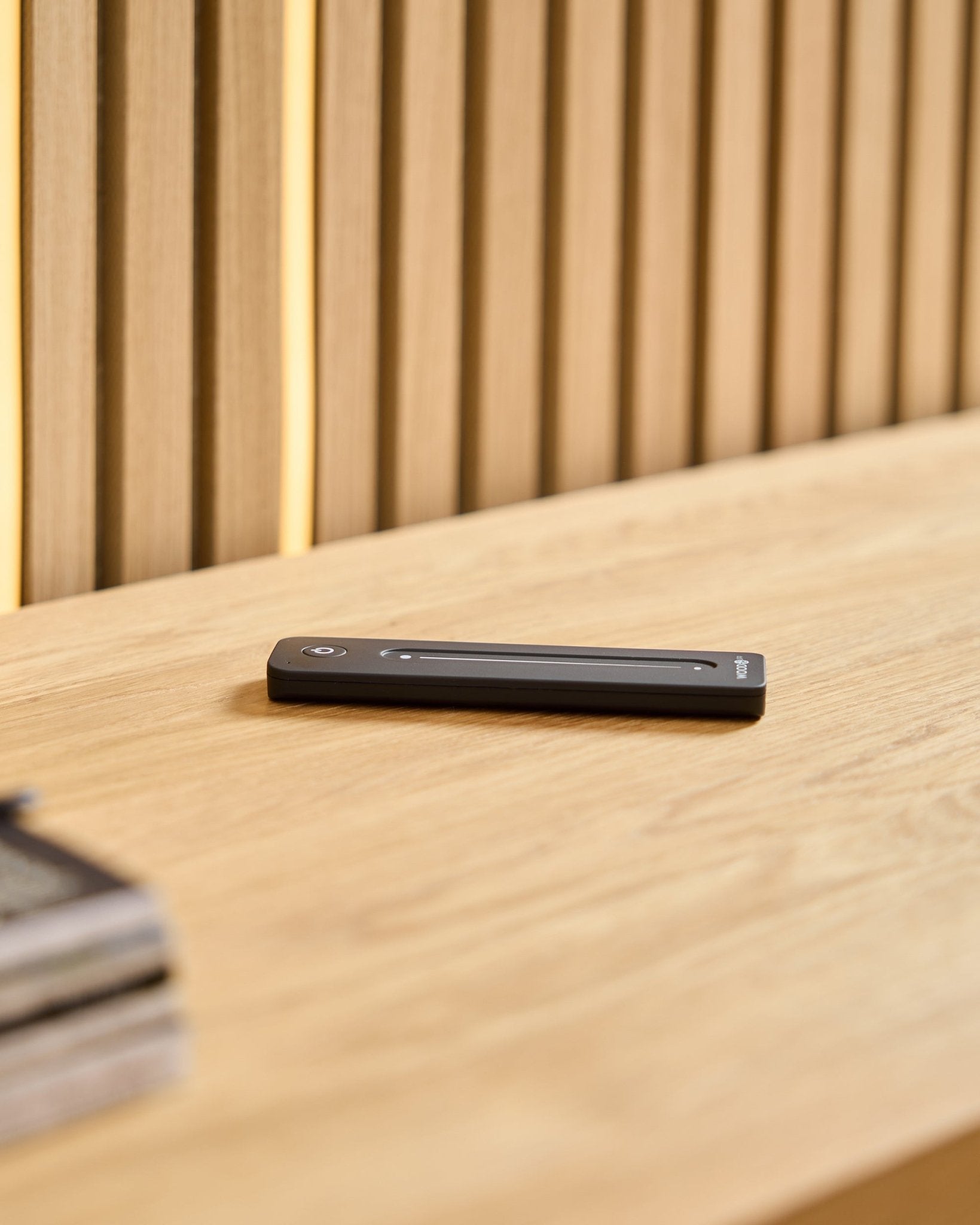 Une télécommande LED WoodUpp black Lumi posée sur une surface en bois clair, sur fond de panneaux acoustiques en bois à lattes.