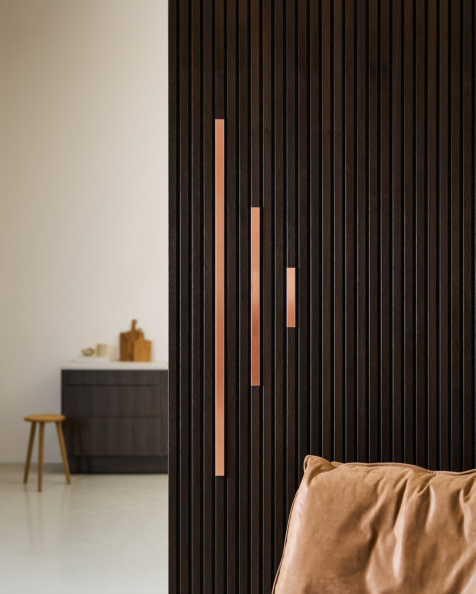 Minimalistischer Raum mit einer dunklen Akustik-Raumteilerwand aus Holzlamellen, WoodUpp Liva Cover in copper, 60-cm-Paneelen mit copper-Griffen, einem hellbraunen Ledersessel und einem Schrankhocker.