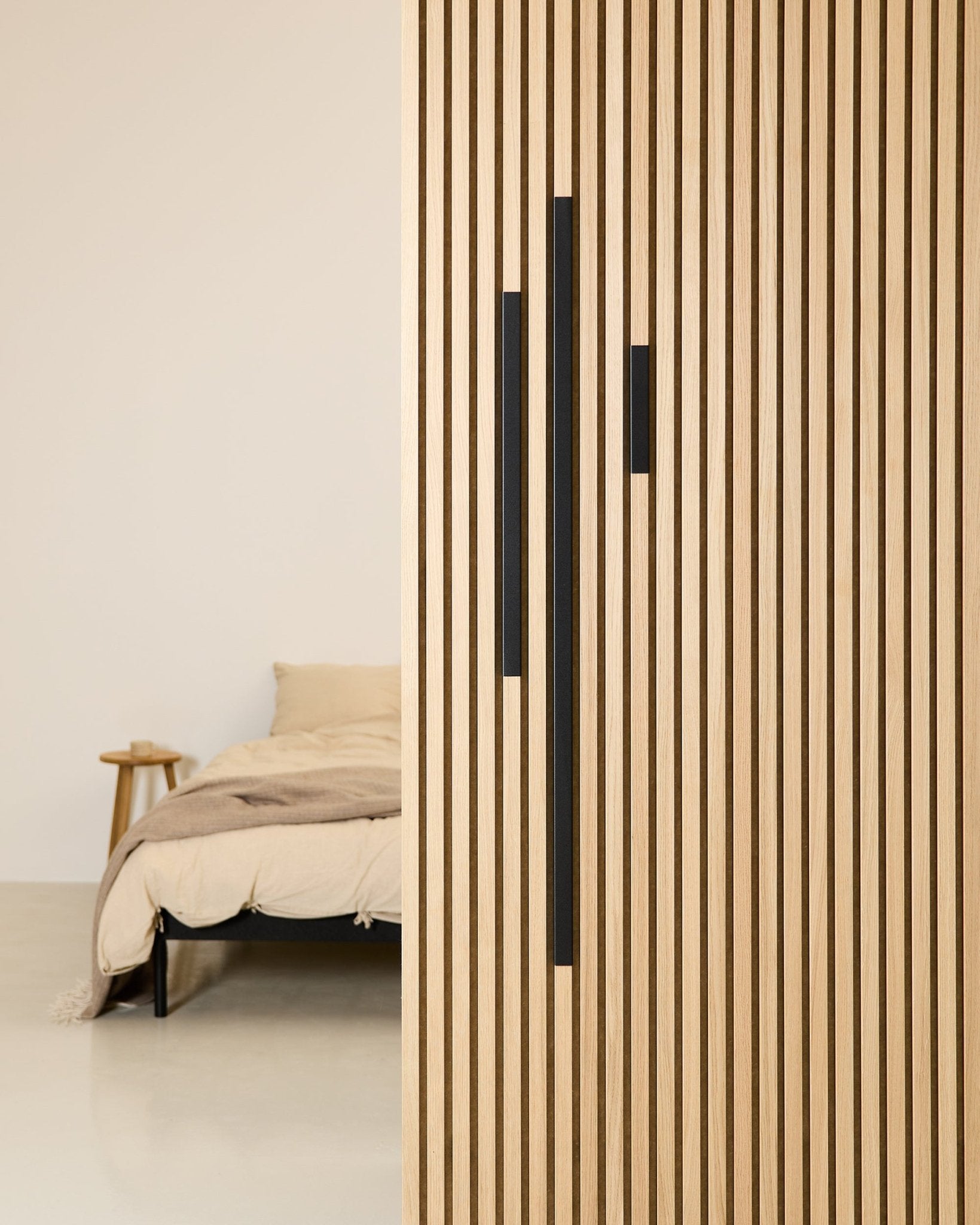 Minimalistisches Schlafzimmer mit einem Bett und einer Decke in beige, teilweise verdeckt durch eine 60 cm breite WoodUpp Akupanel-Trennwand aus Black Aluminum aus der Liva Cover Serie.