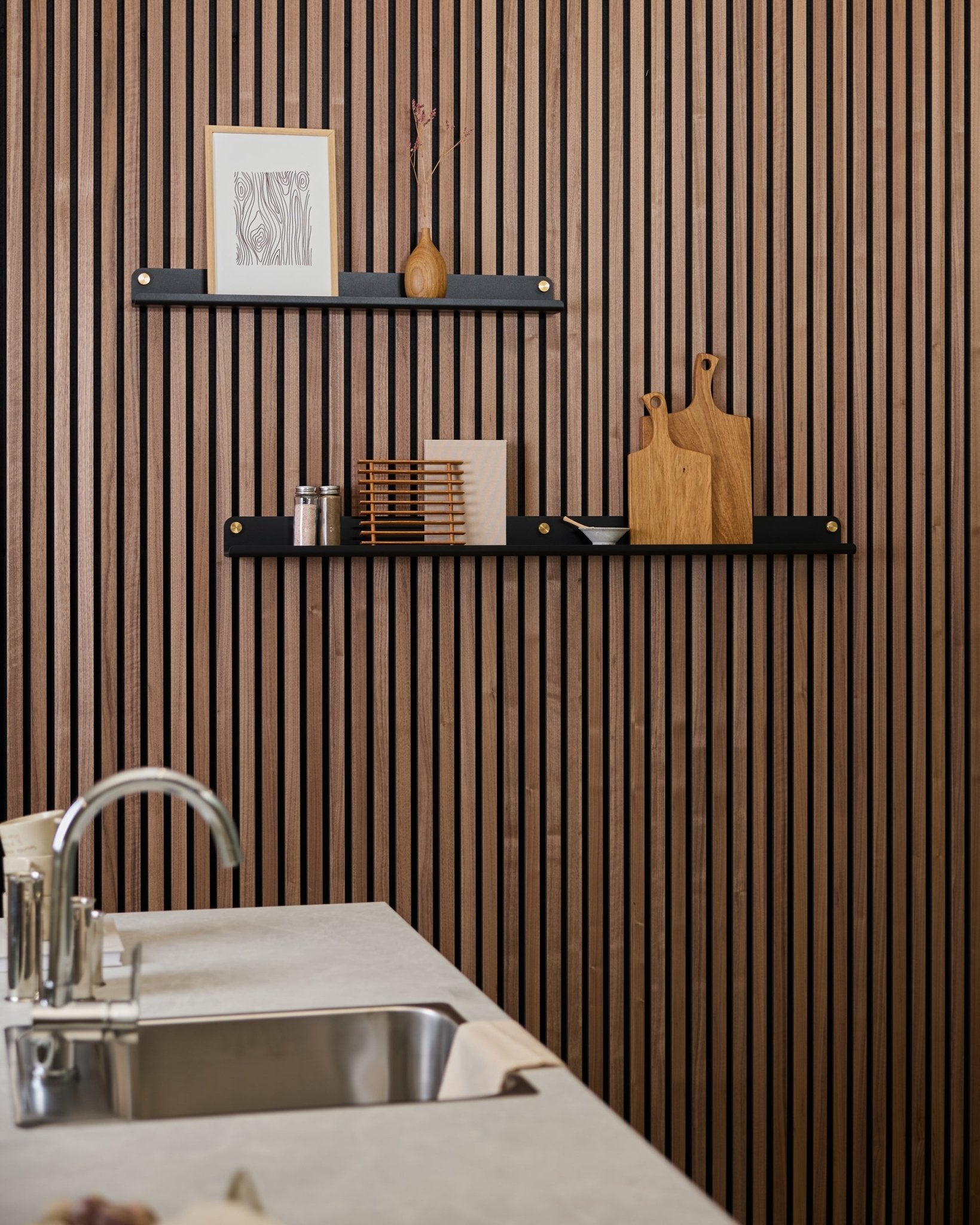 Minimalistische Küche mit einer verticalen Akupanel Akustikwand aus Holzlamellen, blacken Regalen, Schneidebrettern, smallen Deko-Objekten und einer modernen Spültheke davor.