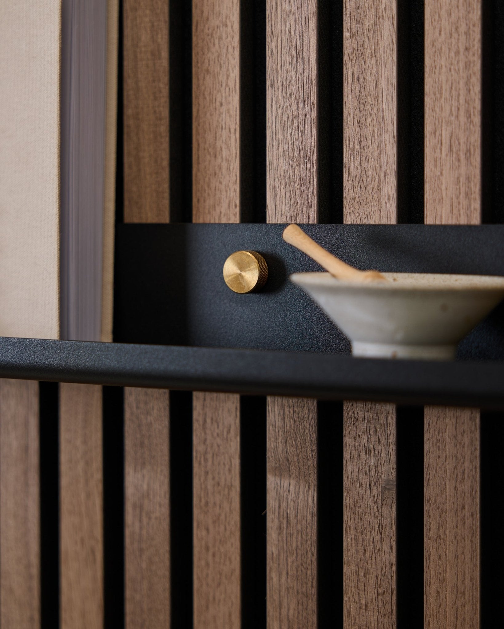 Gros plan sur une étagère étroite WoodUpp black Norr de 115,9 cm, montée sur un panneau acoustique à lattes nordique avec des vis moletées en brass, un bouton doré et un bol en céramique avec cuillère.