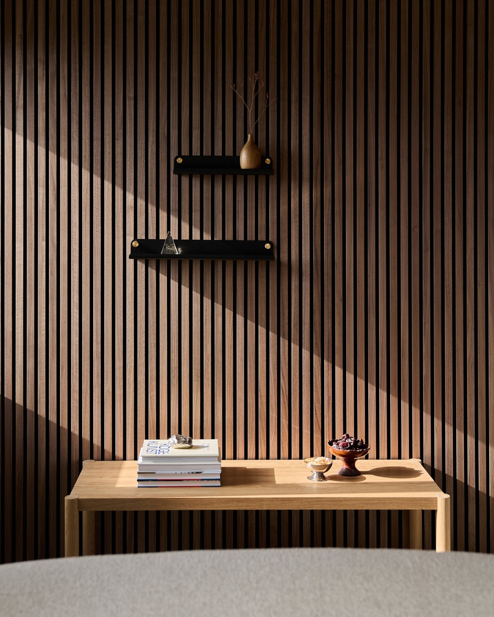 Une table en bois minimaliste avec des livres et des bols est placée près d’un Panneau acoustique à lattes nordique, mettant en valeur l’étagère étroite WoodUpp Norr en black (51,9 cm) ; des ombres baignées de soleil au-dessus.