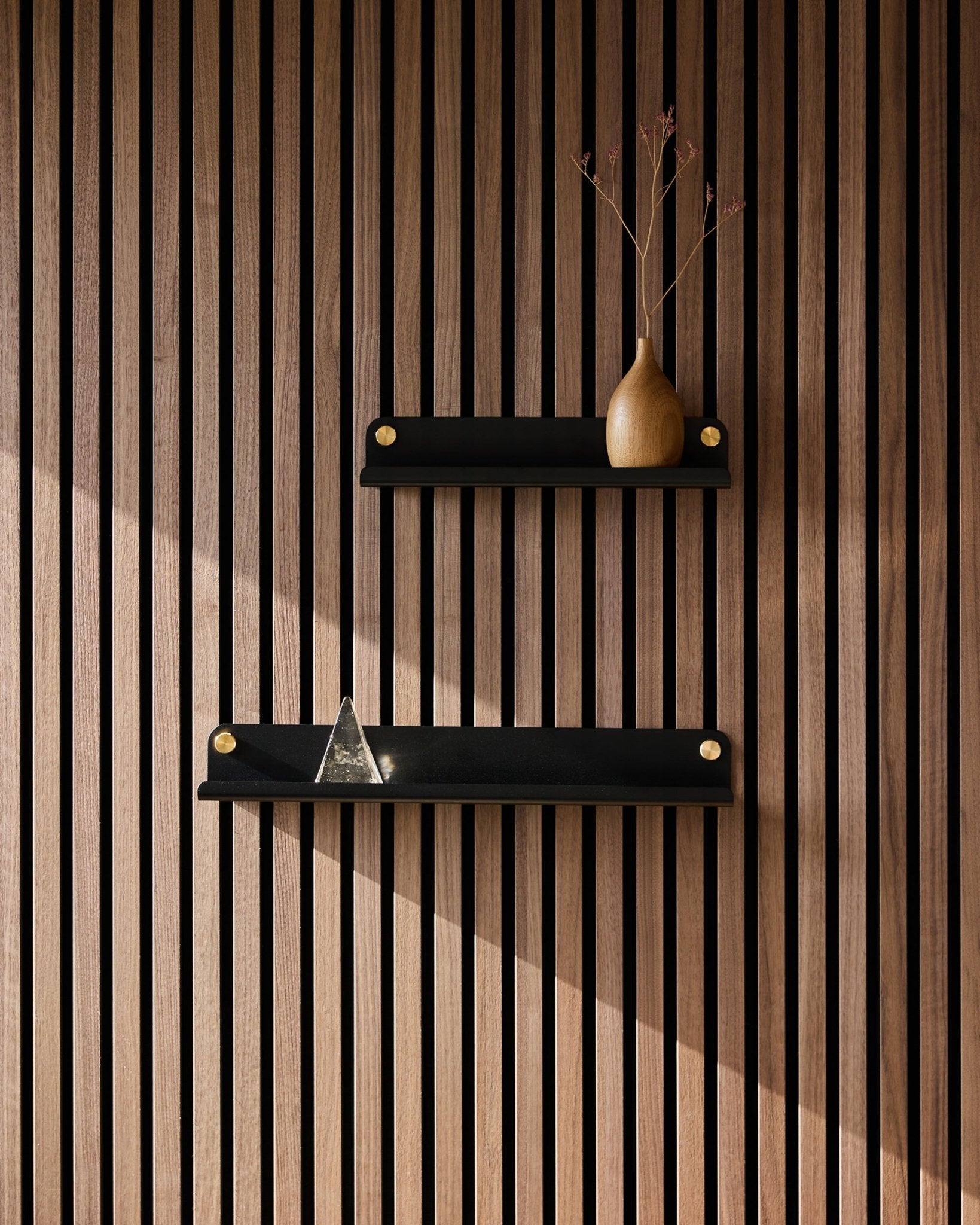 Deux étagères étroites WoodUpp Norr Narrow Shelf en black (35,9 cm), montées sur un panneau acoustique à lattes au style nordique ; celle du haut avec un vase de branches séchées, celle du bas avec une pyramide en verre.