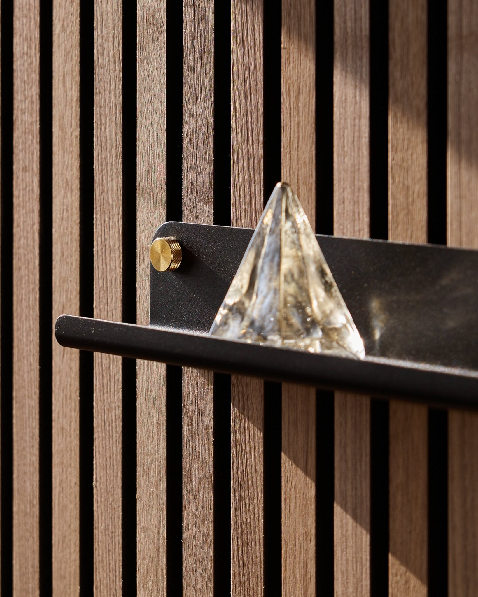 Une pyramide en verre est posée sur l’étagère étroite WoodUpp Norr en black (51,9 cm), montée sur un vertical Panneau acoustique et mise en valeur par la lumière du soleil.