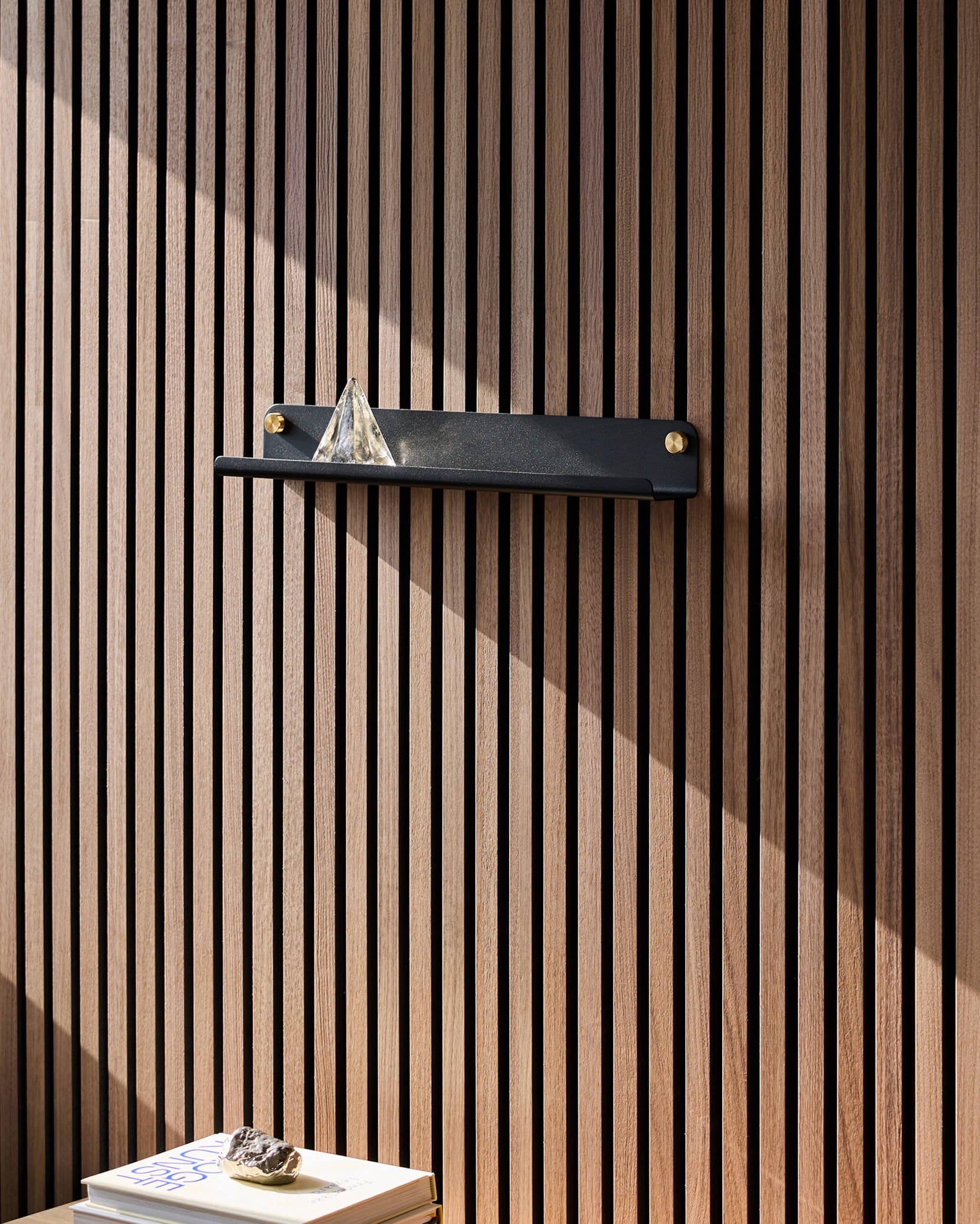 Auf einem schwarzen Norr Narrow Shelf von WoodUpp, das auf einer Akupanel-Akustikholzplatte steht, befindet sich eine Glaspyramide; das Sonnenlicht fällt auf die darunter liegenden Bücher, auf denen ein Stein liegt.