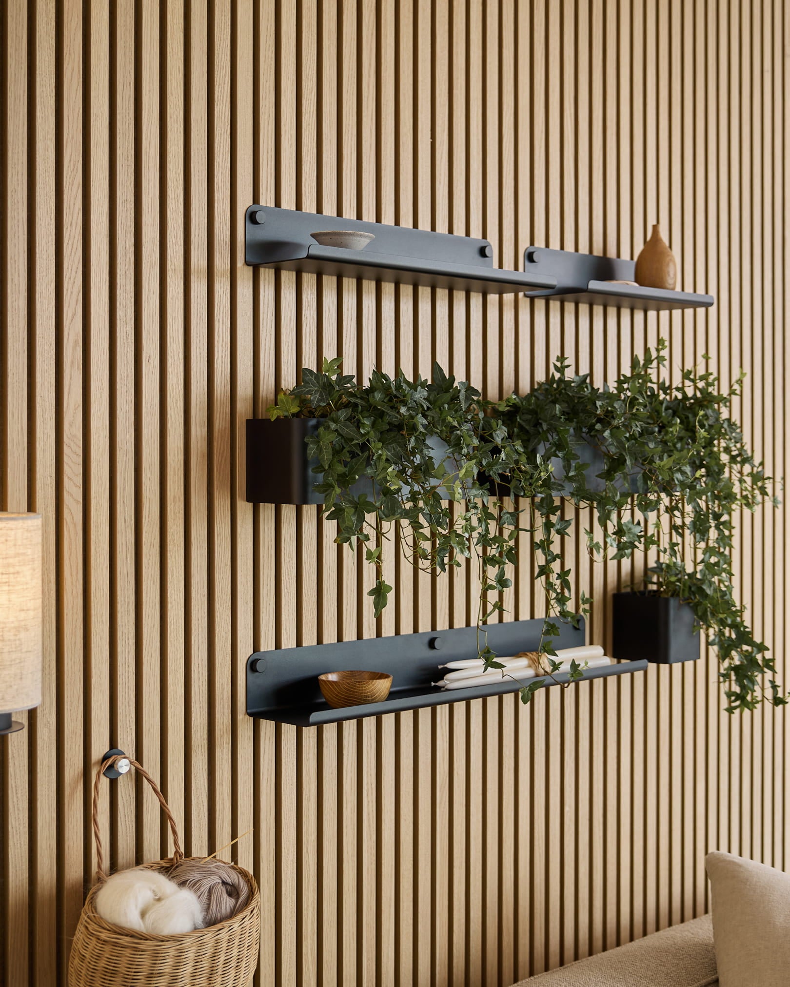 Trois étagères WoodUpp Norr | Plant Box black, avec des objets déco et une plante, sont fixées au-dessus d’un panier en osier rempli de pelotes, sur un panneau acoustique mural à lattes vert.