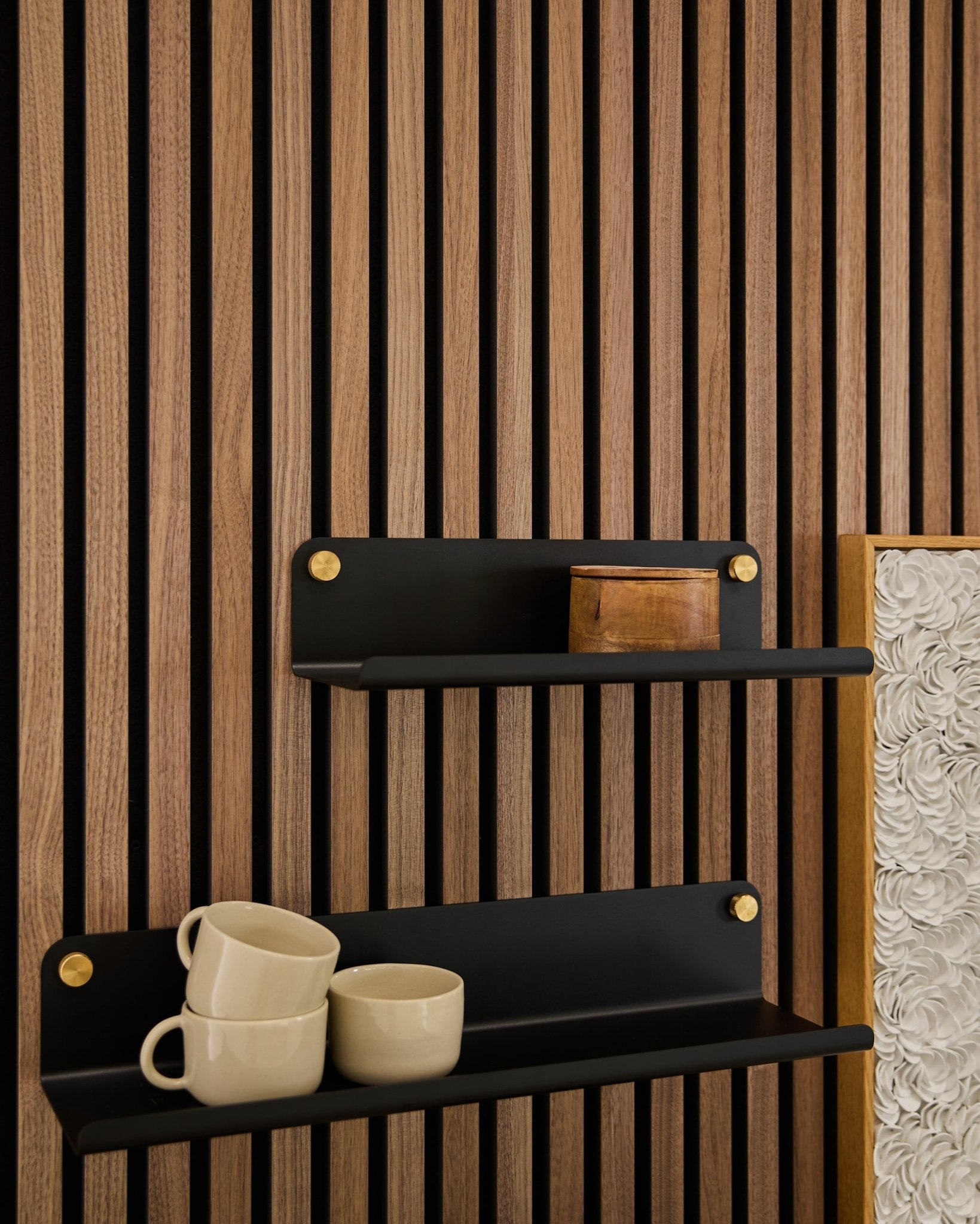 Deux étagères larges black Norr | Wide Shelves de WoodUpp (35,9 cm) avec des supports dorés, montées sur un panneau acoustique en bois Akupanel ; l’étagère du bas accueille 3 mugs beige, celle du haut une boîte en bois small.