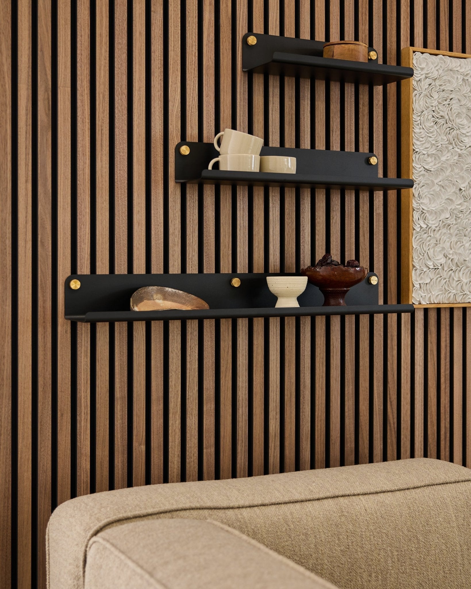 Drei blacke Regalböden, darunter das WoodUpp Norr Wide Shelf (75,9 cm), präsentieren Deko auf einer Holzlatten-Akustikpaneel-Wand über einem beigefarbenen Sofa; ein Teil des Kunstwerks ist sichtbar.