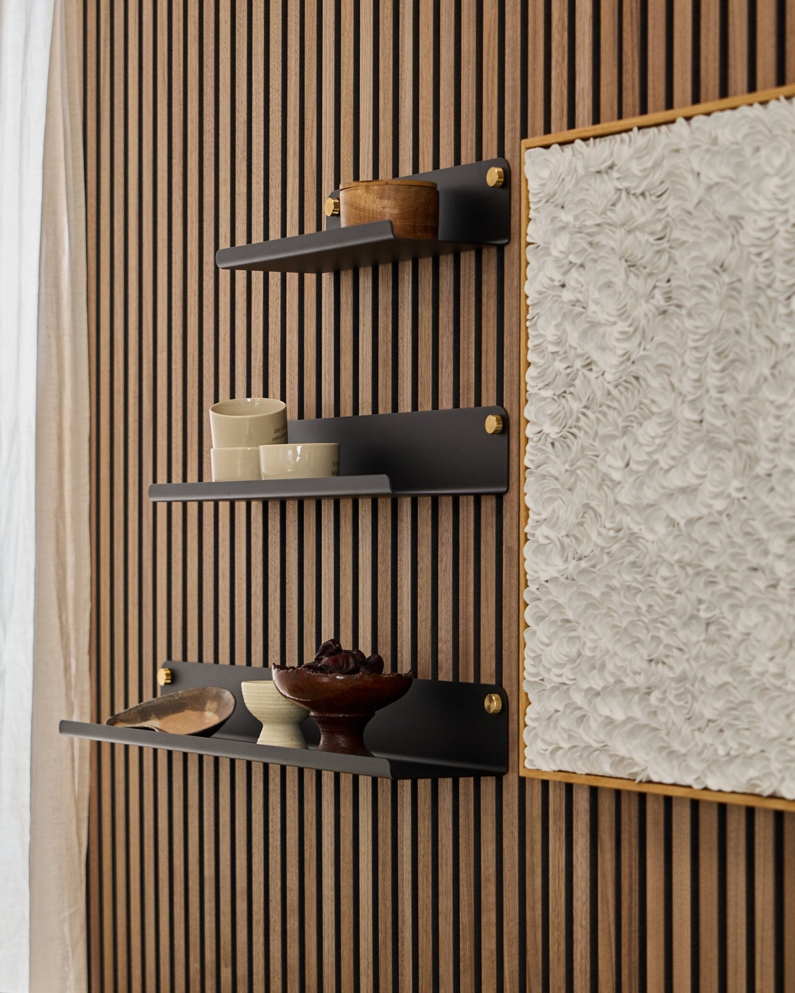 Trois étagères larges WoodUpp Norr de 51,9 cm en black, montées sur un panneau acoustique en bois WoodUpp Akupanel, à côté d’une œuvre d’art blanche texturée, avec des bols et des contenants en bois exposés dessus.