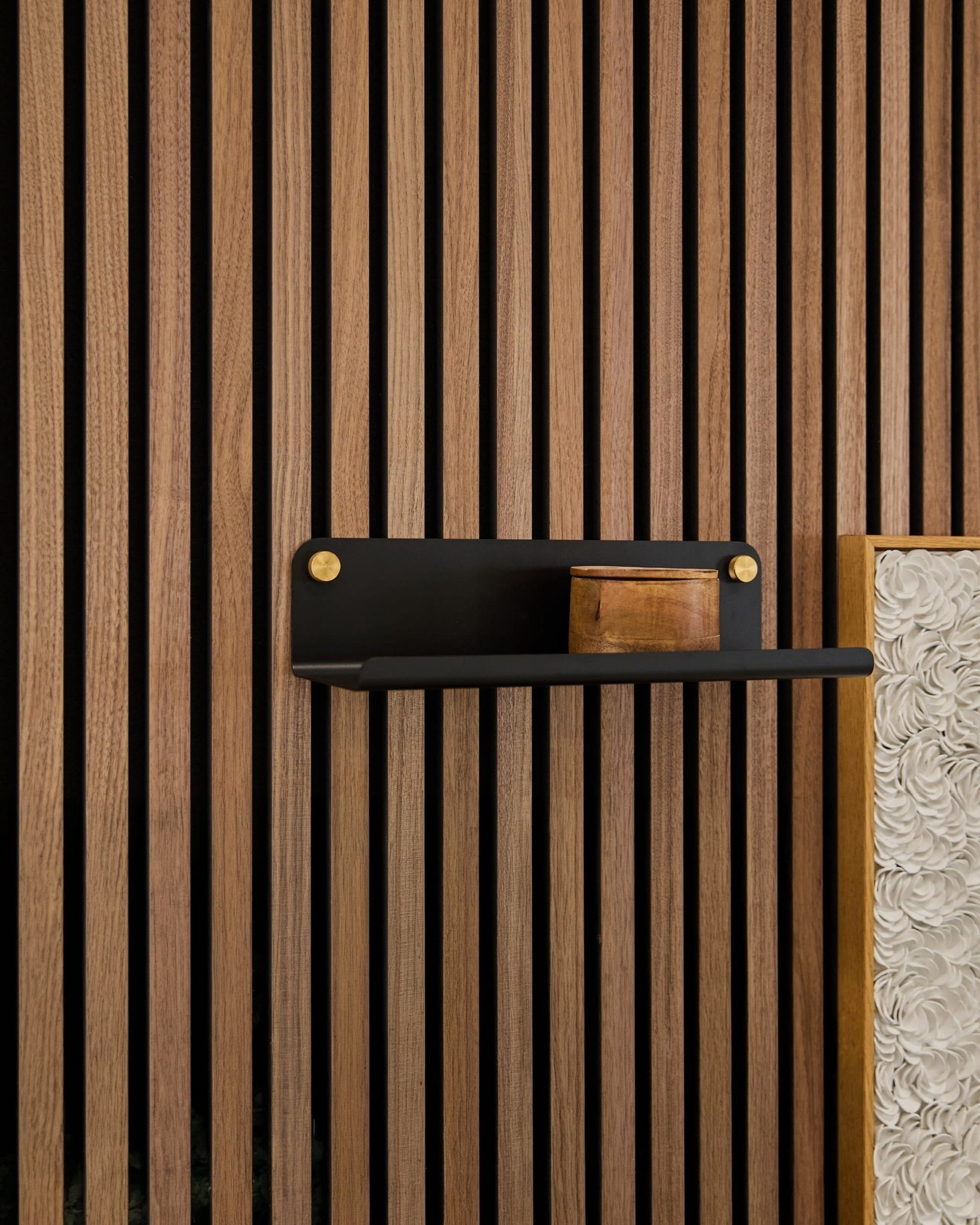 Une petite boîte en bois posée sur une étagère WoodUpp Norr Wide (noire, 35,9 cm) fixée contre des lattes acoustiques verticales en bois Akupanel, avec un panneau blanc texturé visible.