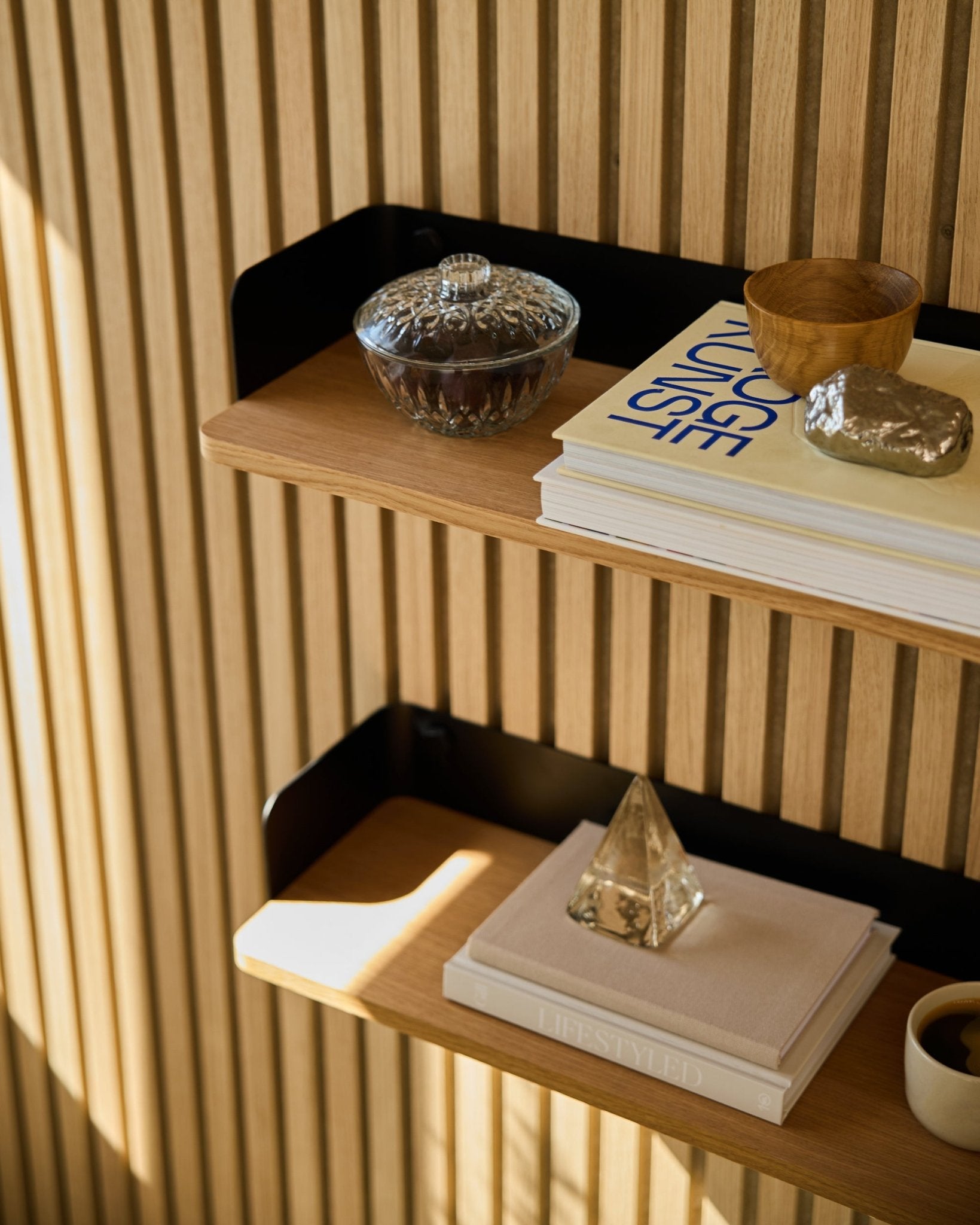 La bibliothèque WoodUpp Linn Bookshelf en Natural Oak (55,9 cm) accueille un bocal en verre, une tasse, une pyramide dorée et des livres, devant des panneaux acoustiques en bois Akupanel baignés de soleil.