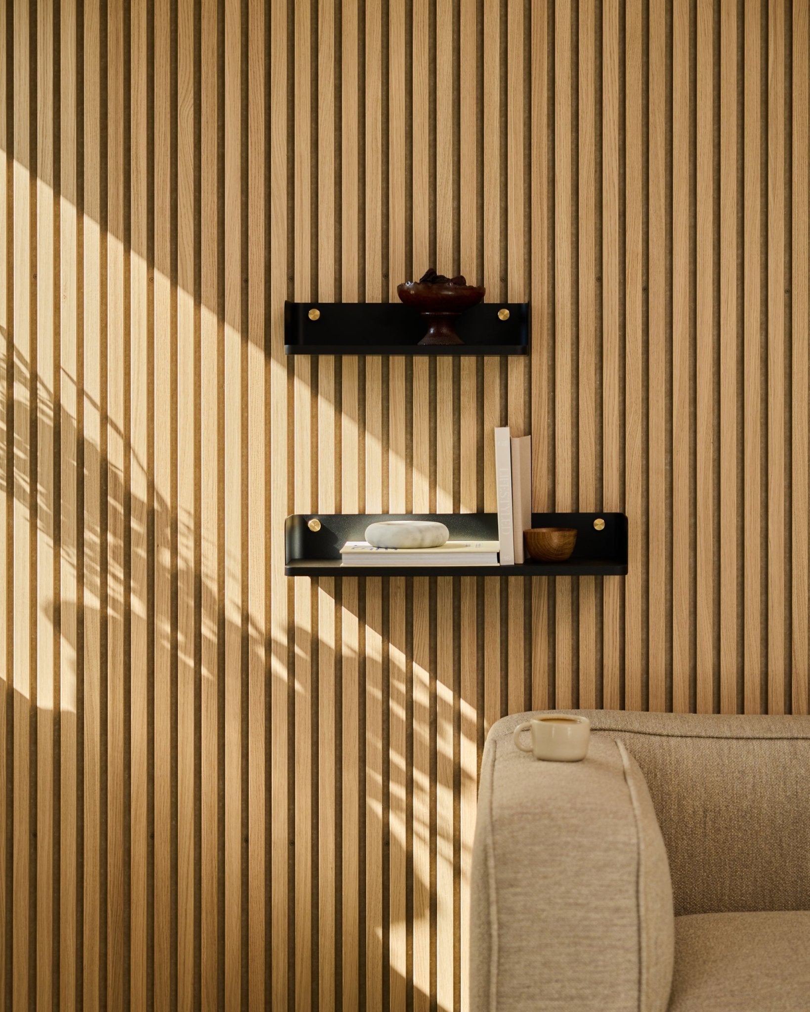 La lumière du soleil éclaire un canapé beige et l’étagère Linn de WoodUpps en black oak, 55,9 cm, installés devant un Panneau acoustique minimaliste de style nordique à lattes.