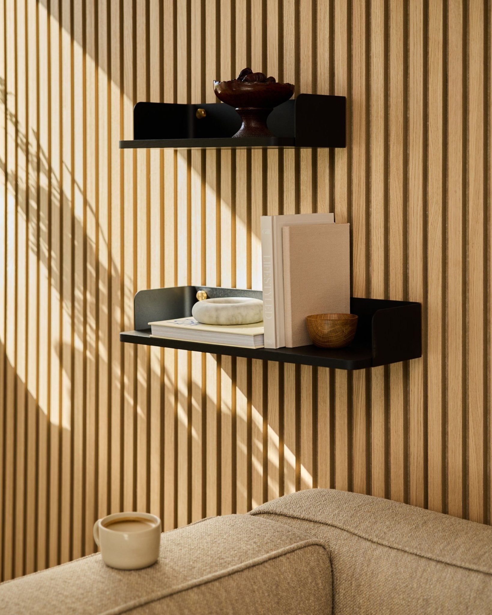 Une étagère WoodUpp en black oak (55,9 cm) présente des livres, des objets déco et un bol sur un mur en panneaux de bois acoustiques ; un canapé beige et une tasse au premier plan.