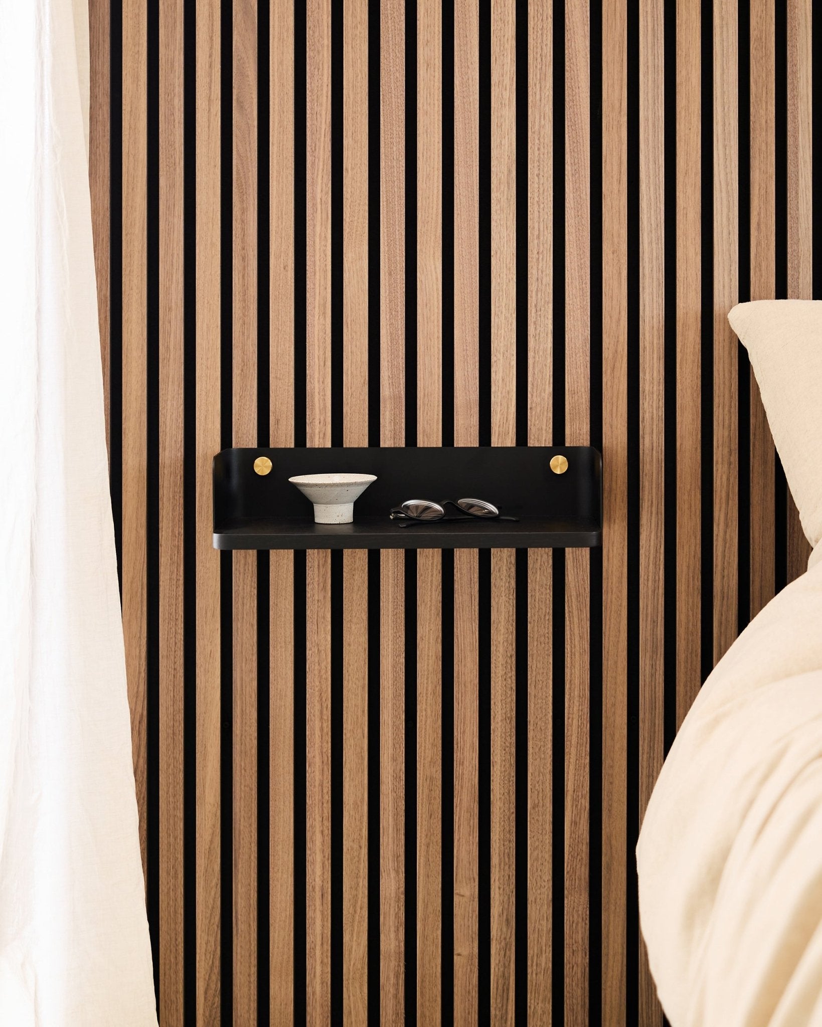 L’étagère Linn en Black Oak de WoodUpp est fixée sur un panneau acoustique à lattes au style nordique, et accueille un bol et des verres à côté d’un lit avec du linge de lit beige.