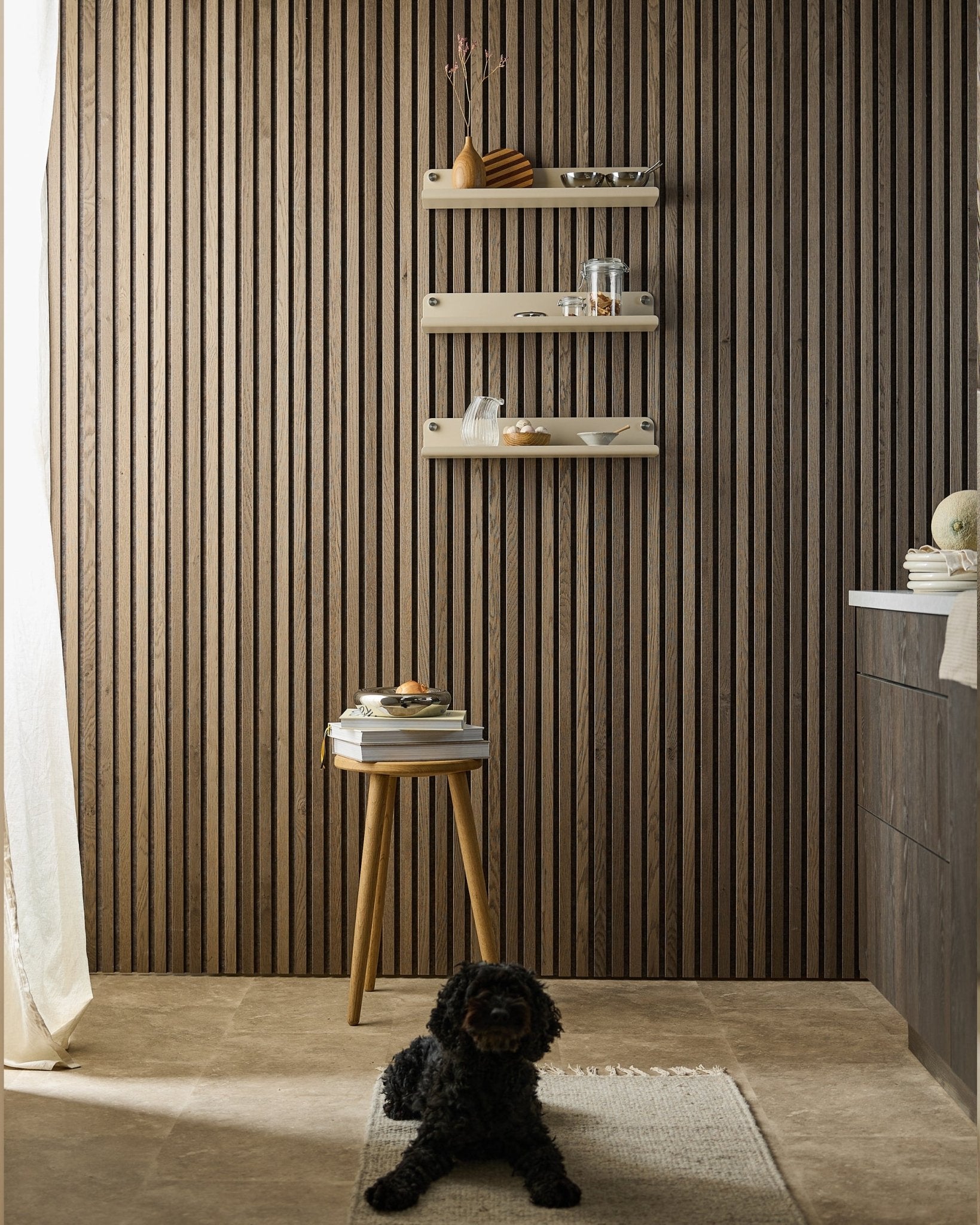 Ein blacker Hund liegt auf einem Teppich in einem modernen Raum mit einer Akupanel Akustikpaneel Wand, einem beigen WoodUpp Norr Wide Shelf (51,9 cm), einem Hocker mit Büchern und weichem Licht.