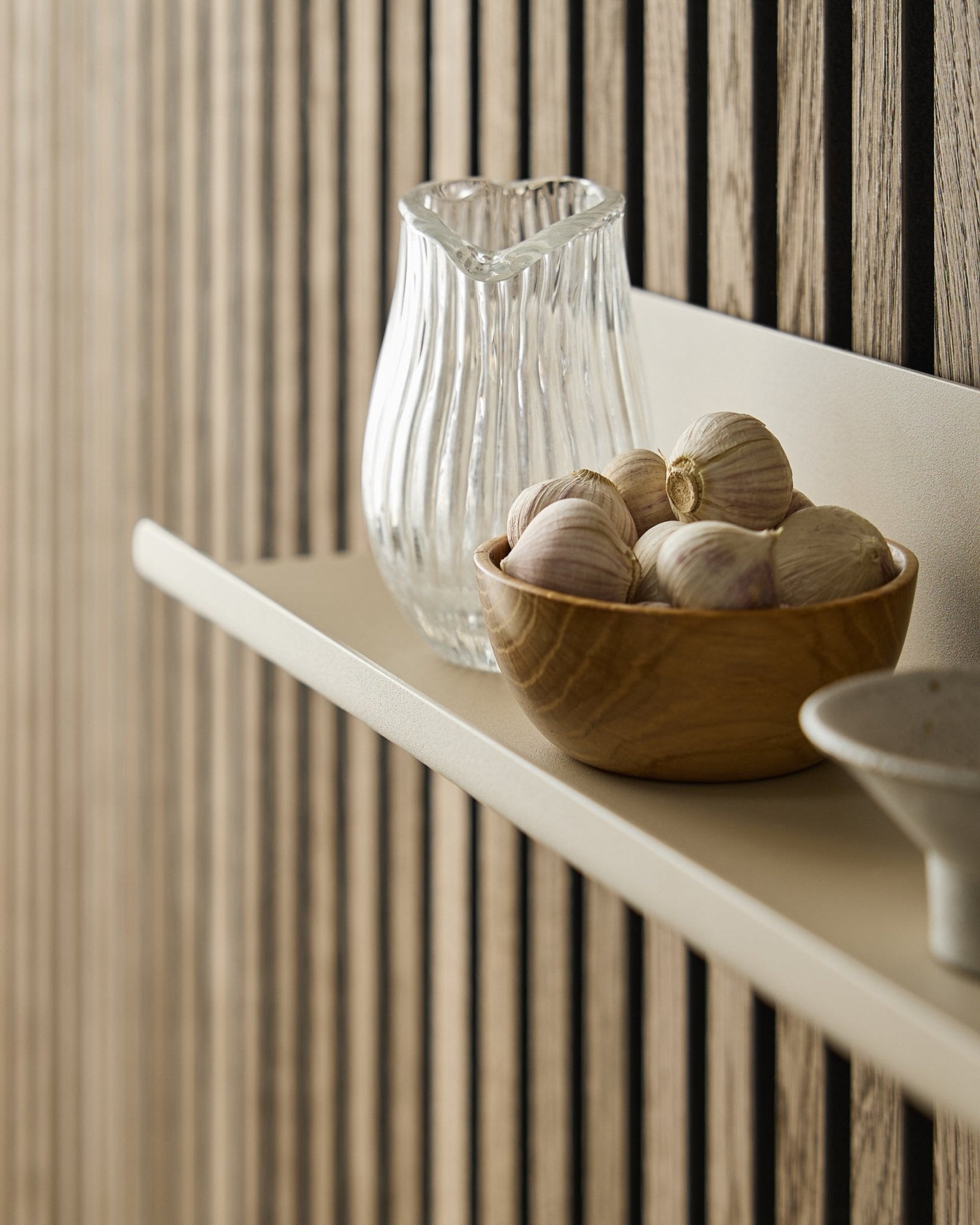 Un bol en bois rempli d’ail et une carafe en verre sur l’étagère large Norr de WoodUpp, fixée sur un Akupanel Panneau acoustique avec des lattes en bois verticales.