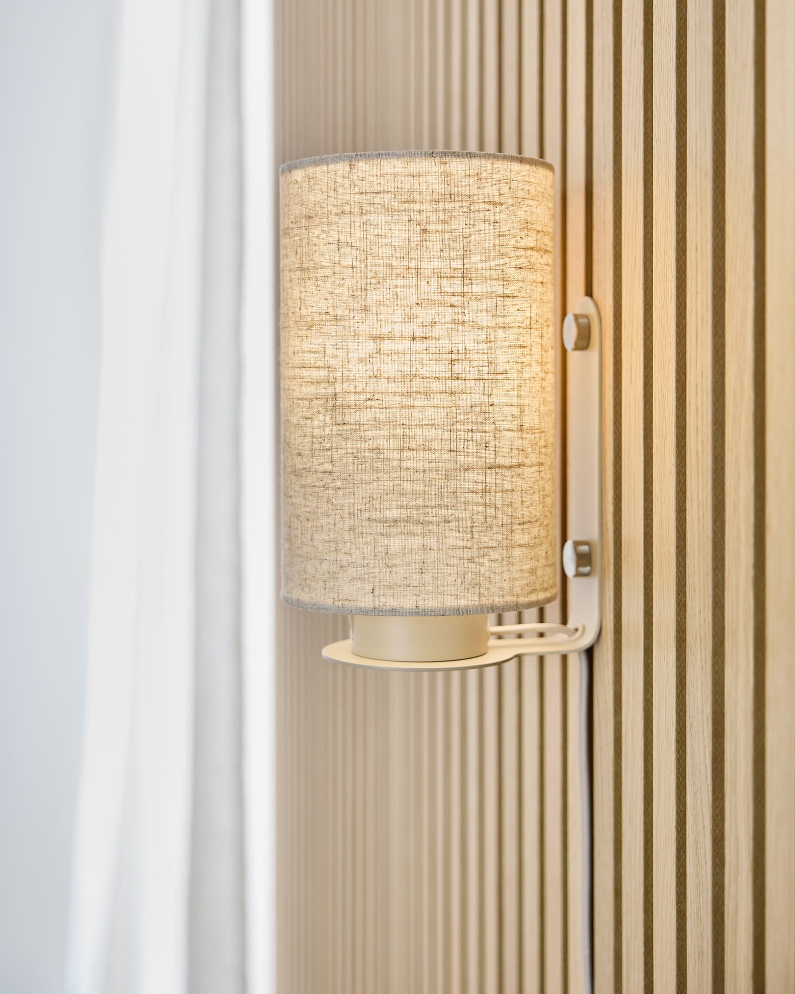 La lampe murale Feya en Beige de WoodUpp diffuse une lumière chaleureuse sur un mur acoustique à lames beige en Akupanel, placée à côté d’un rideau blanc et d’un abat-jour en lin-coton.