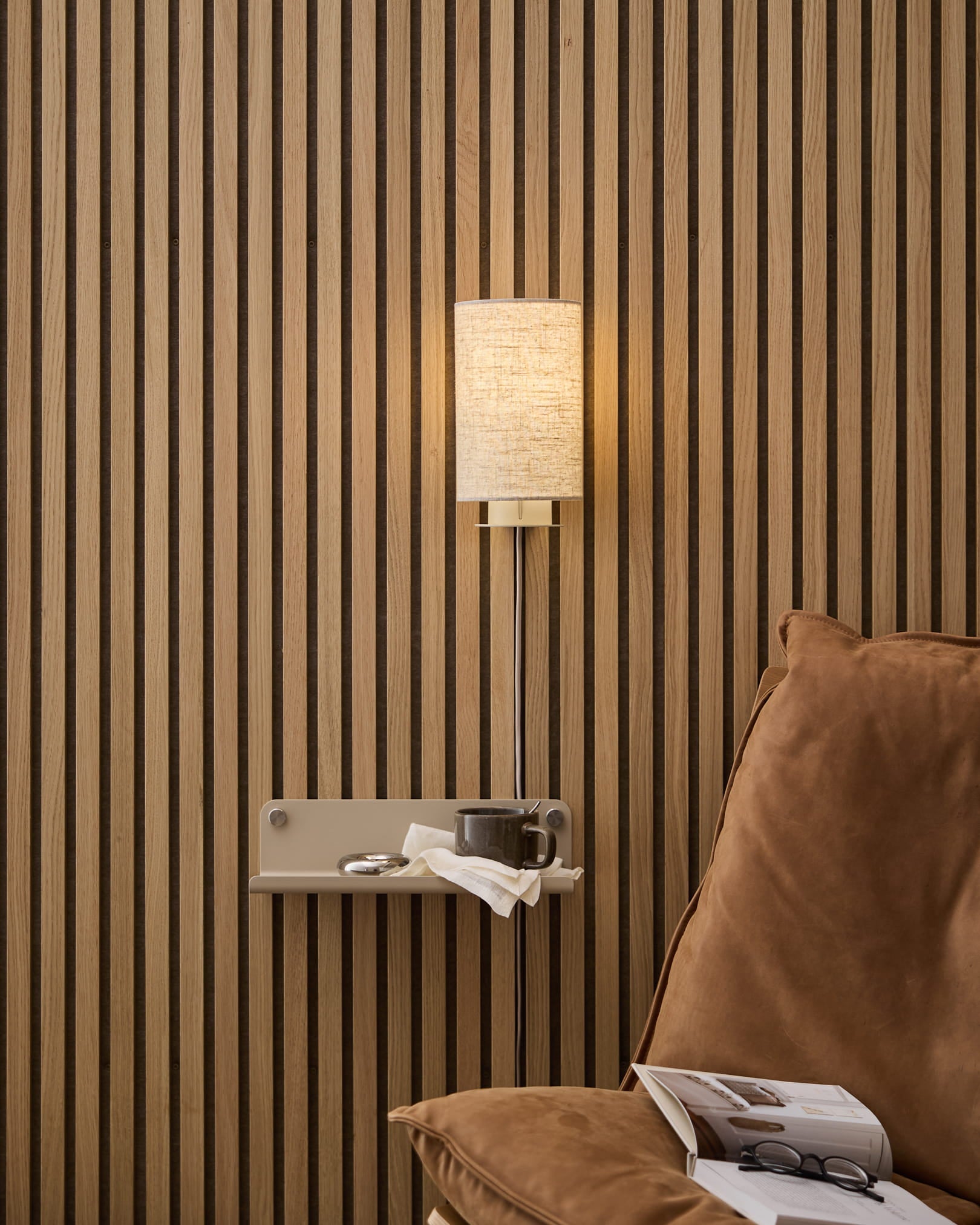 Eine beige Feya Wall Lamp aus Leinen-Baumwolle von WoodUpp, montiert über einem gemütlichen Sessel und einem Beistelltisch mit Buch und Tasse, vor einem nordischen Holzlamellen-Akustikpaneel.