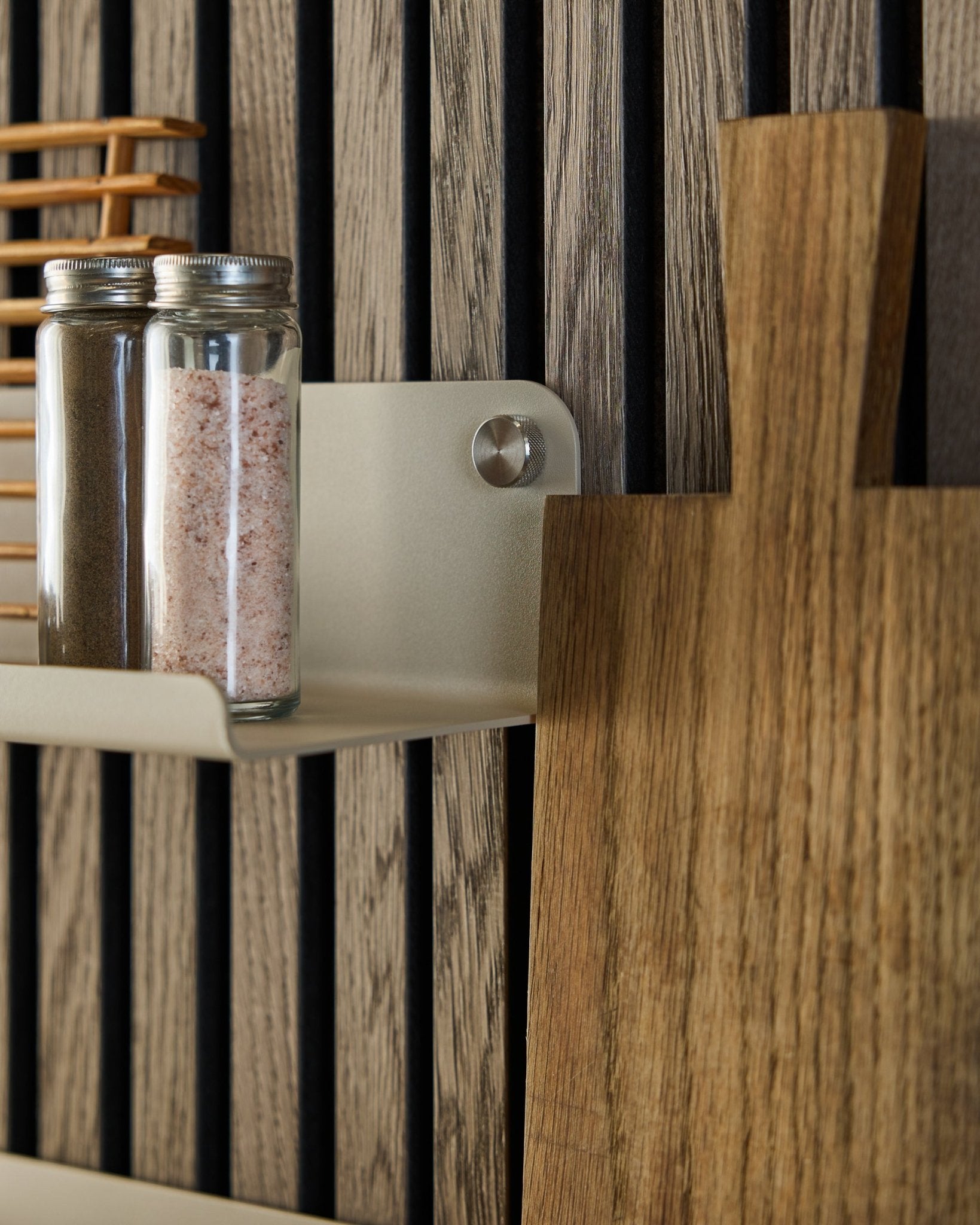 Des bocaux en verre contenant des épices et une planche à découper en bois reposent sur l’étagère large WoodUpp Norr en beige, 35,9 cm, fixée sur un panneau acoustique à lattes vertical.