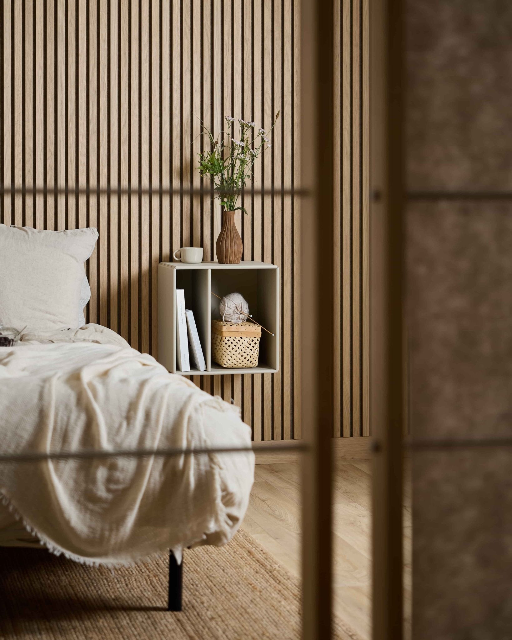 Chambre à coucher minimaliste avec la bibliothèque WoodUpp Arka de couleur beige, des livres, un panier et un vase à fleurs, le tout disposé devant un panneau acoustique à lamelles Akupanel de style nordique.