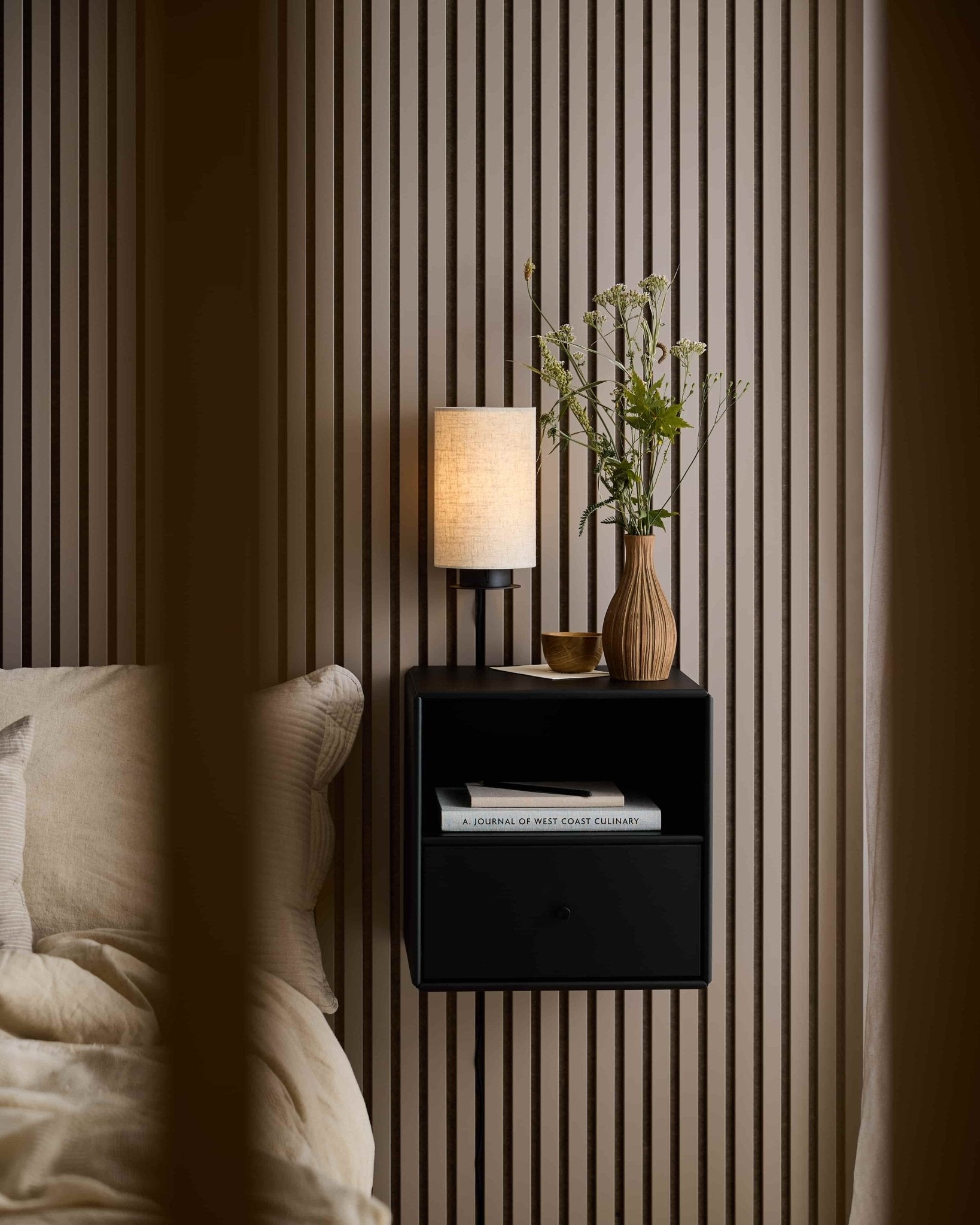 Table de chevet avec lampe, vase, tasse et livre, placée à côté dun lit, ainsi quun panneau acoustique WoodUpp Akupanel de couleur « Dark Cashmere » destiné à labsorption acoustique.