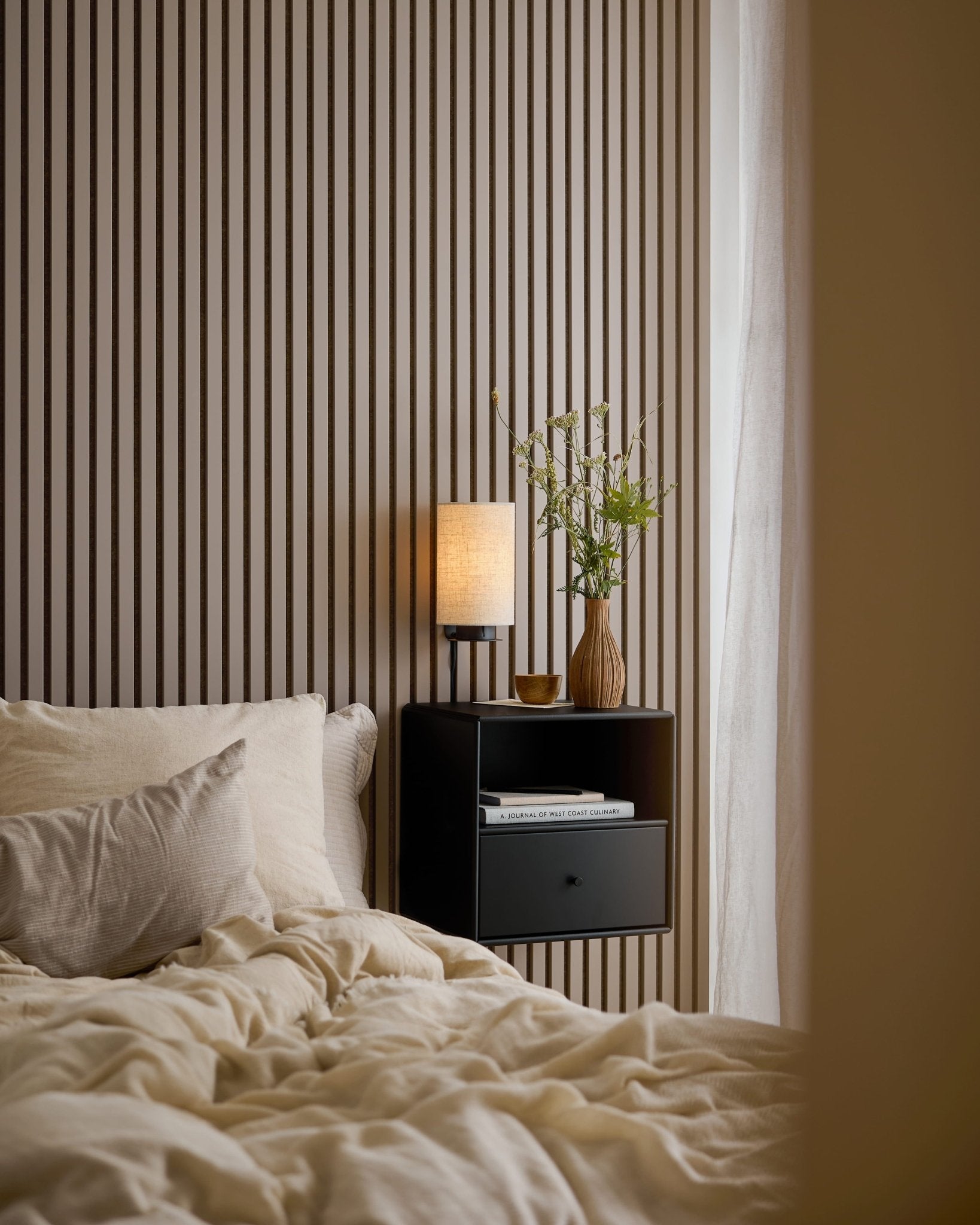 Lit avec une parure de lit couleur crème, des livres et une lampe posée sur une table de chevet noire, à côté dun panneau acoustique WoodUpp Akupanel Dark Cashmere, alliant style et isolation phonique.