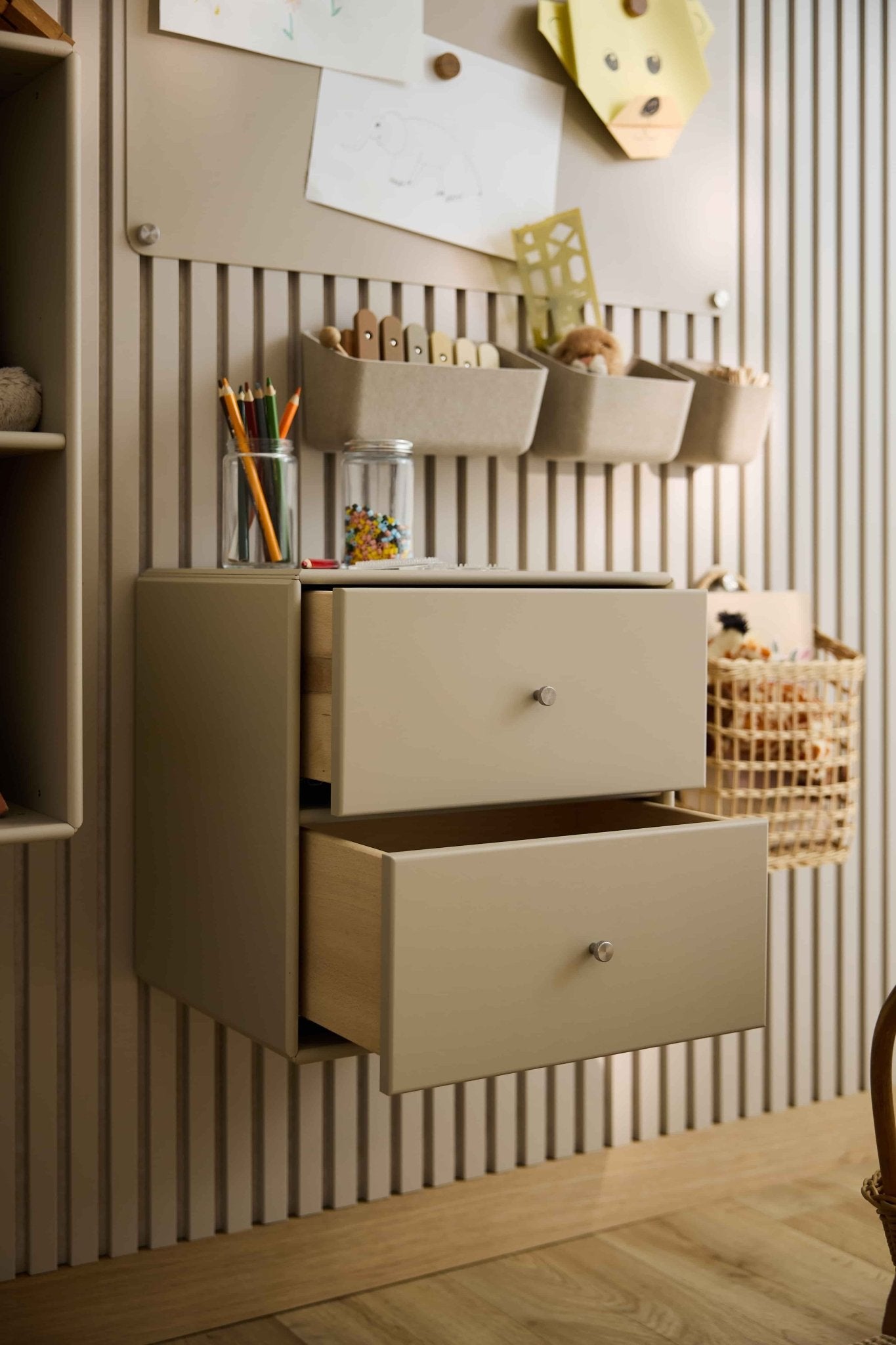 Deux commodes « Arka » de couleur beige, signées WoodUpp, partiellement ouvertes, servent à ranger du matériel artistique et des jouets ; des rangements fermés et des paniers sont disposés à proximité.