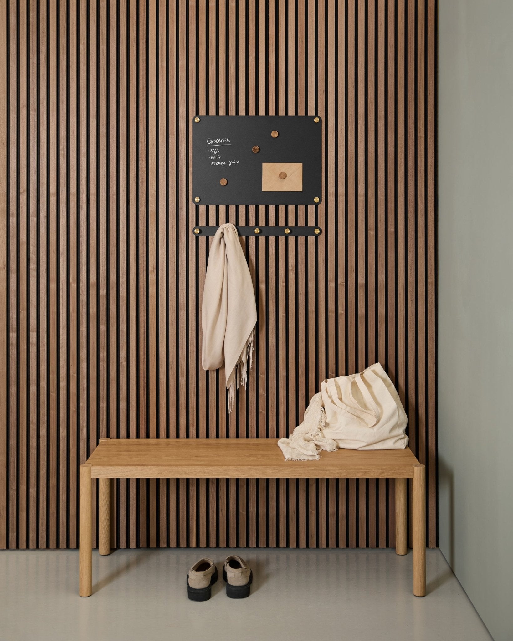 Entrée minimaliste avec un banc en bois, deux écharpes, des sandales et un tableau magnétique WoodUpp small Norr sur un panneau mural acoustique à lattes en bois Akupanel.