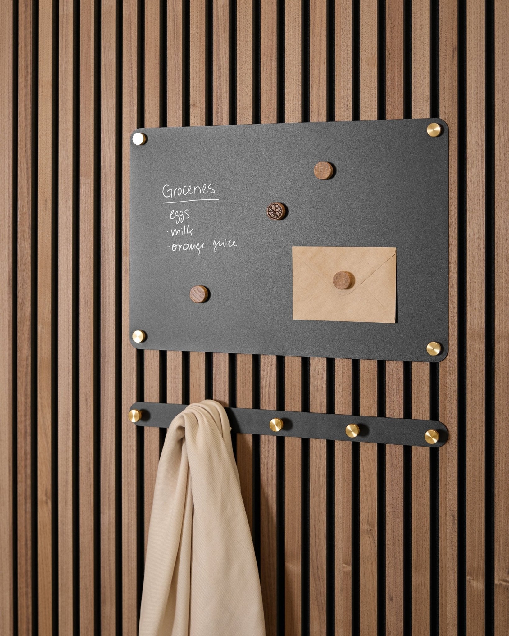 Eine small black Norr Magnettafel von WoodUpp hält eine Einkaufsliste, einen Umschlag, Magnete und ein beiges Tuch an Haken und ist auf einem Akupanel Akustikpaneel montiert.