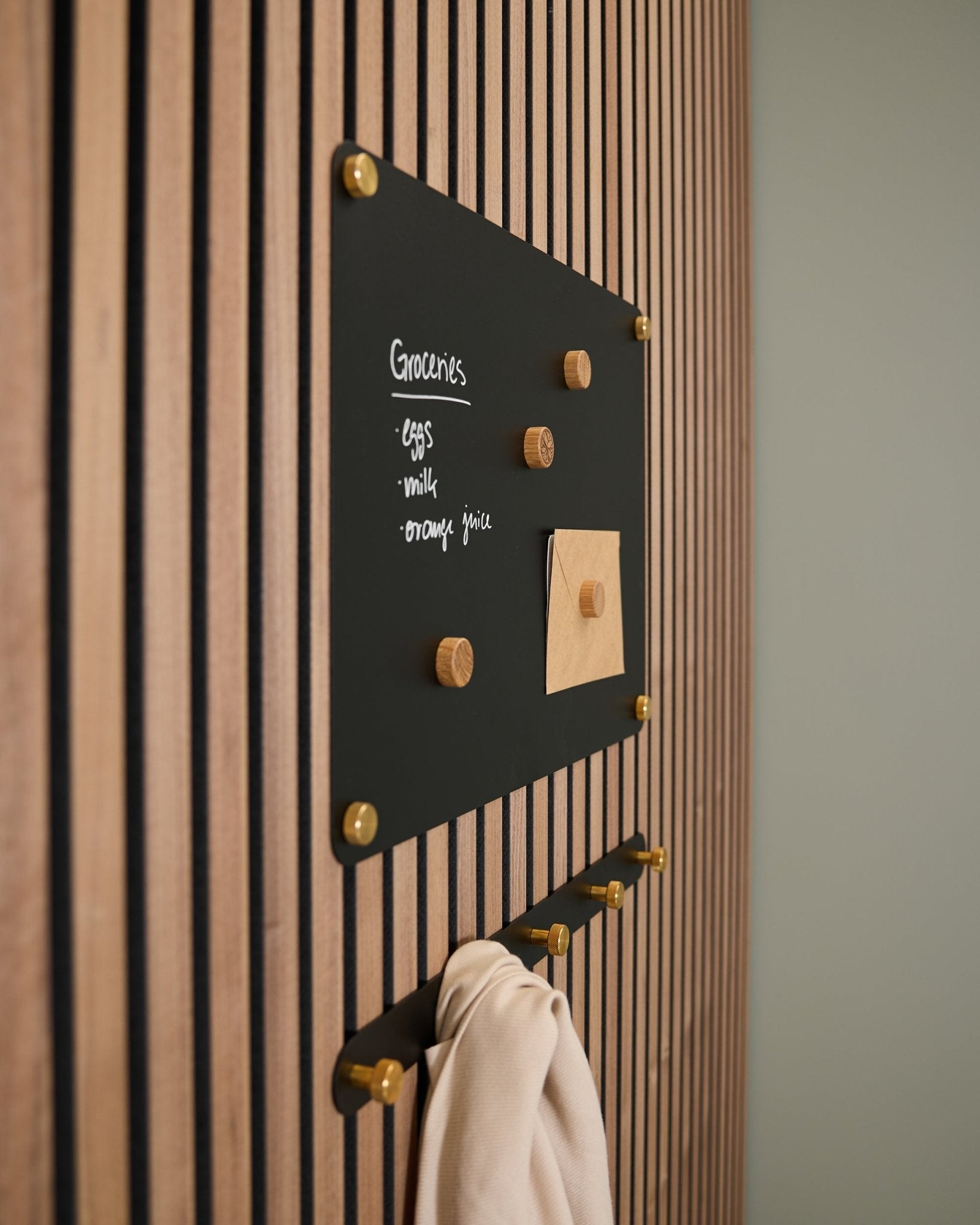 Eine small black Norr Magnettafel von WoodUpp, montiert auf einem Akupanel Akustik-Holzpaneel, zeigt eine Einkaufsliste und eine Notiz; darunter hängen an Haken ein helles Tuch.