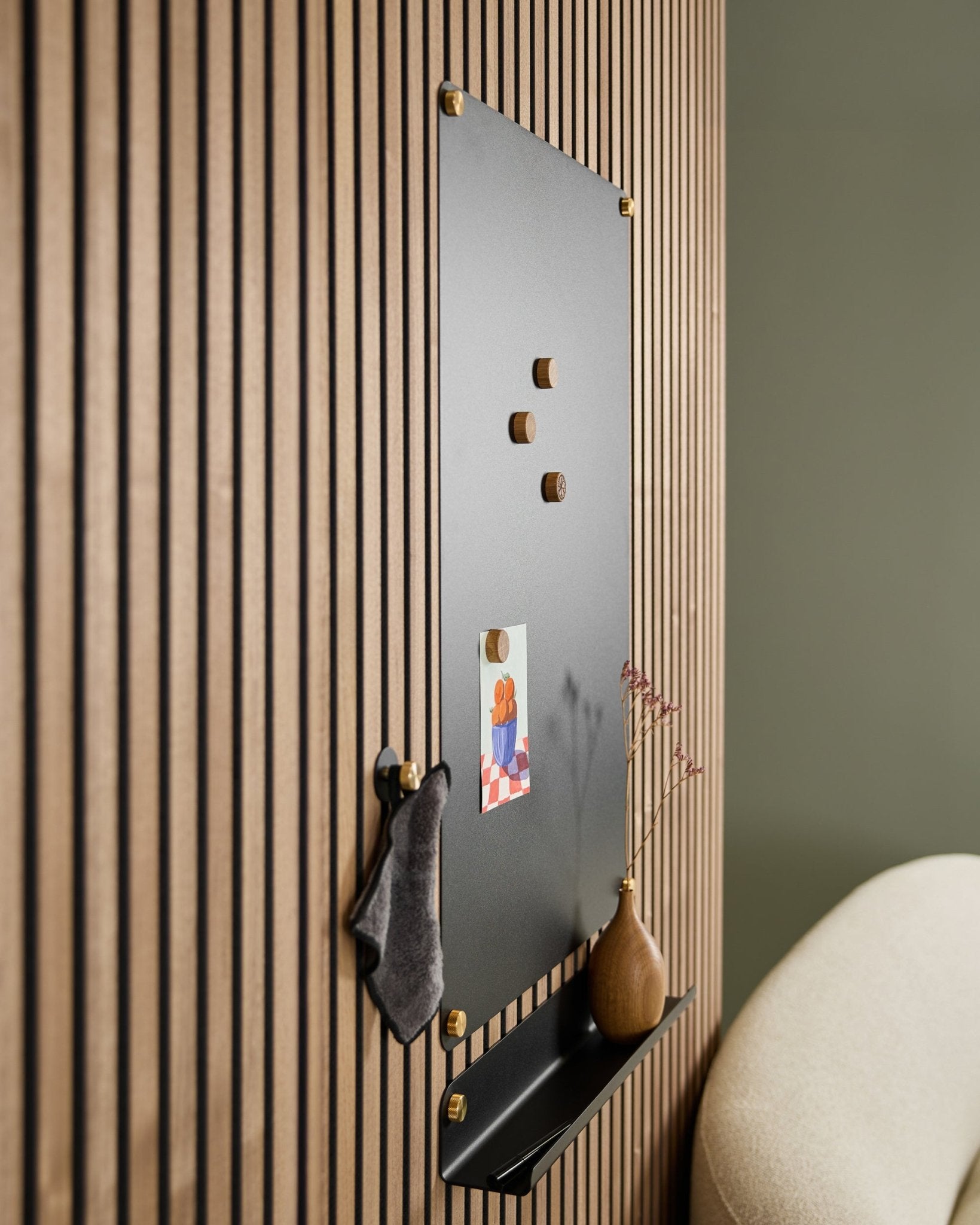 Die WoodUpp Norr large black Akustik-Magnettafel mit Holzmagneten, Pinnwand, Ablage und Vase ist an der Wand neben einem beigen Stuhl montiert.