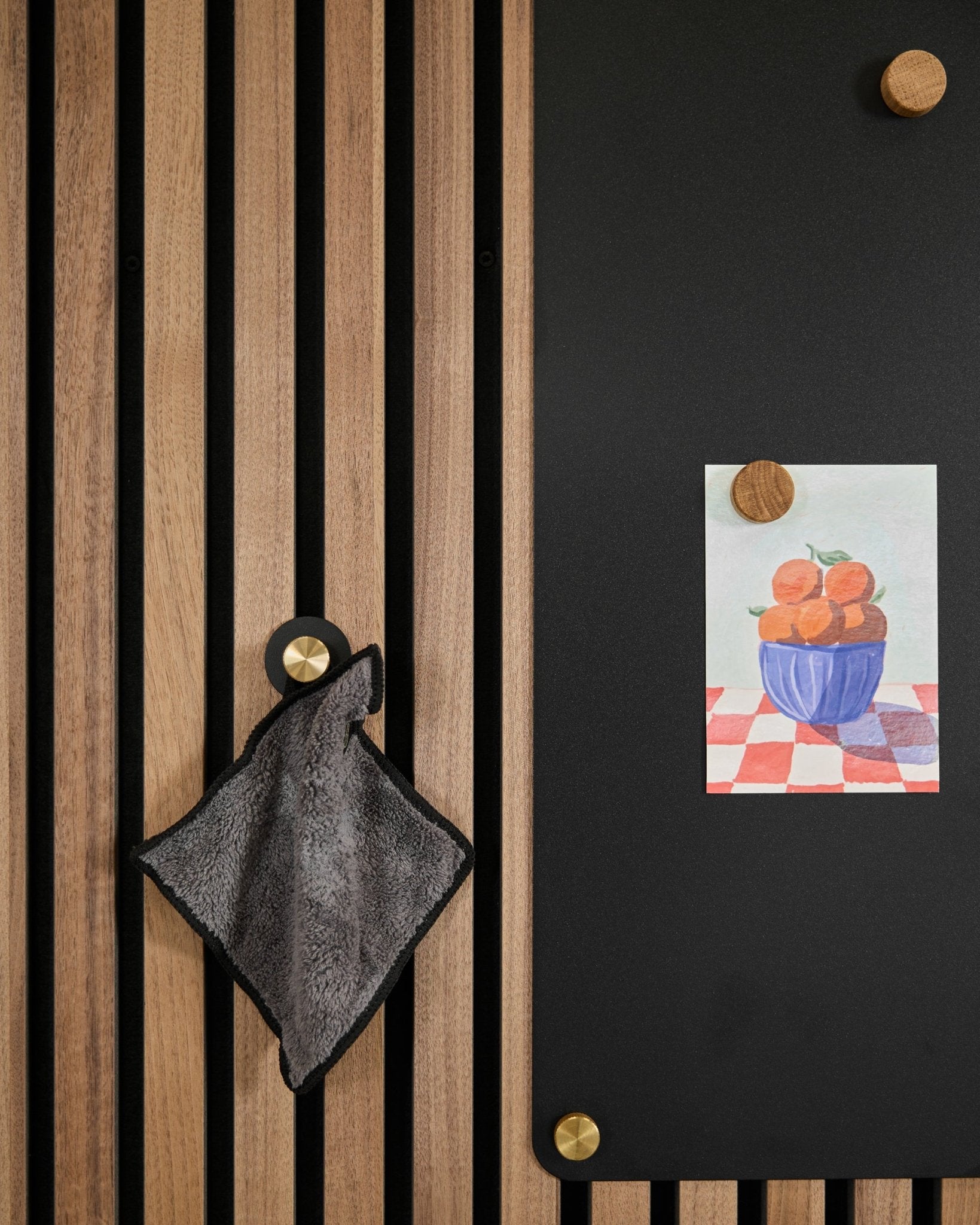 Ein smalles Handtuch hängt an einem nordischen Akustik-Lamellenpaneel neben einer blacken Tafel, auf der eine Zeichnung von Orangen in einer Schale zu sehen ist, befestigt mit zwei goldenen Magneten.