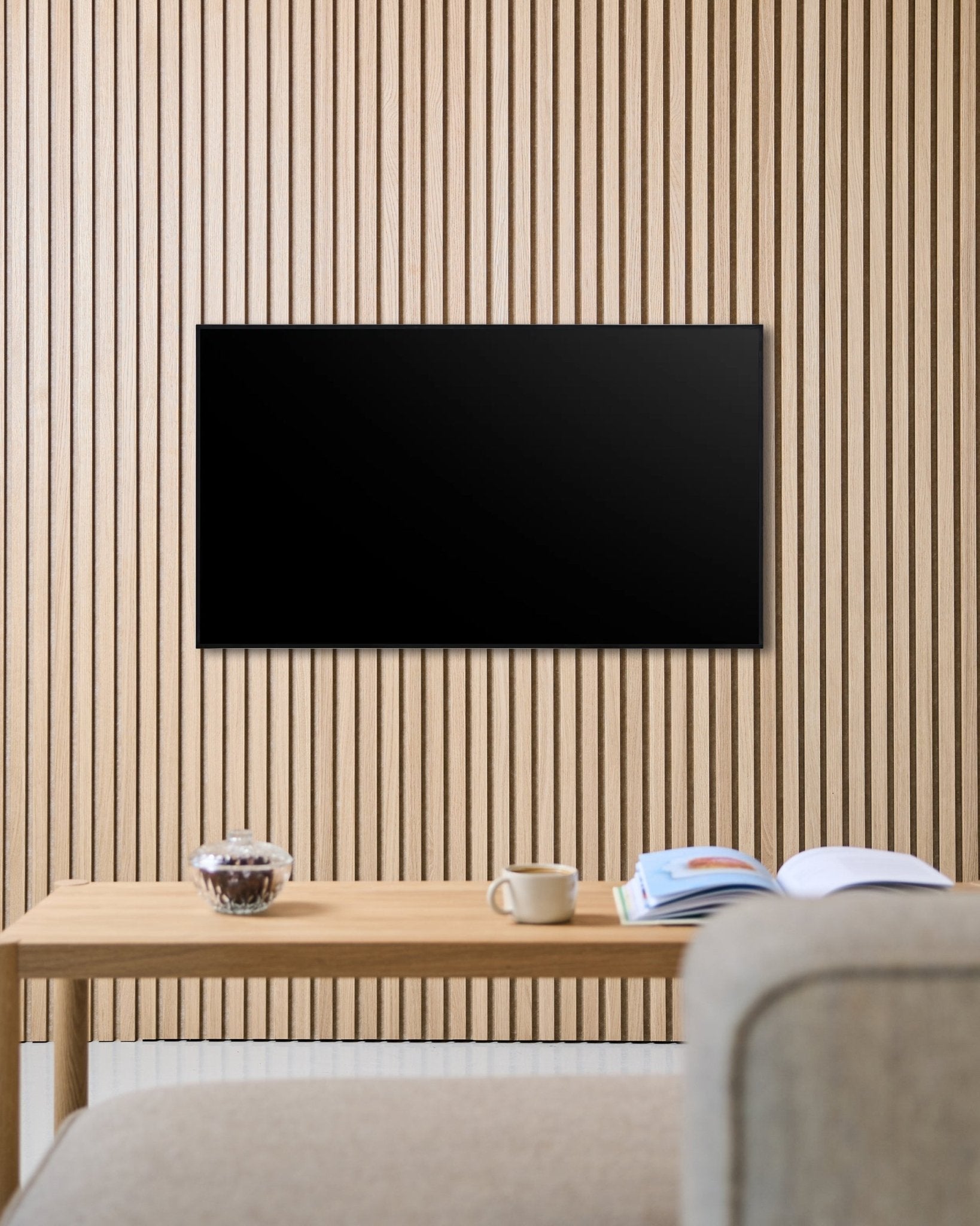 Un support TV Fjell (small) de WoodUpp fixe un écran plat sur un mur acoustique en tasseaux de bois Akupanel au style nordique, au-dessus d’une table avec une tasse, un bol et un magazine ouvert, à côté d’un canapé beige.