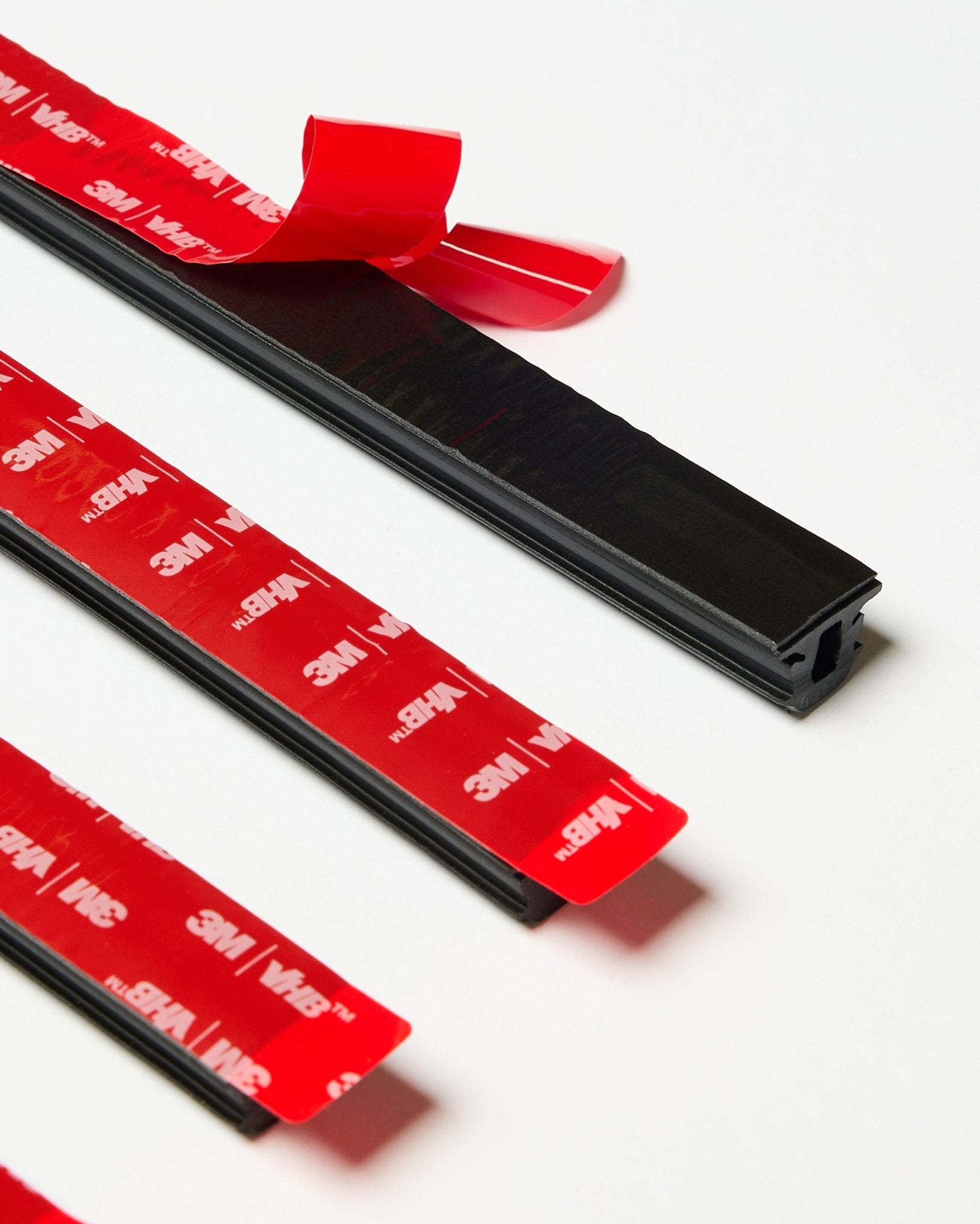 Set aus vier black WoodUpp CREATE Pop-on Profile Leisten mit sichtbarer roter 3M-Klebefolie, eine Leiste teilweise abgezogen, auf einer weißen Oberfläche präsentiert.