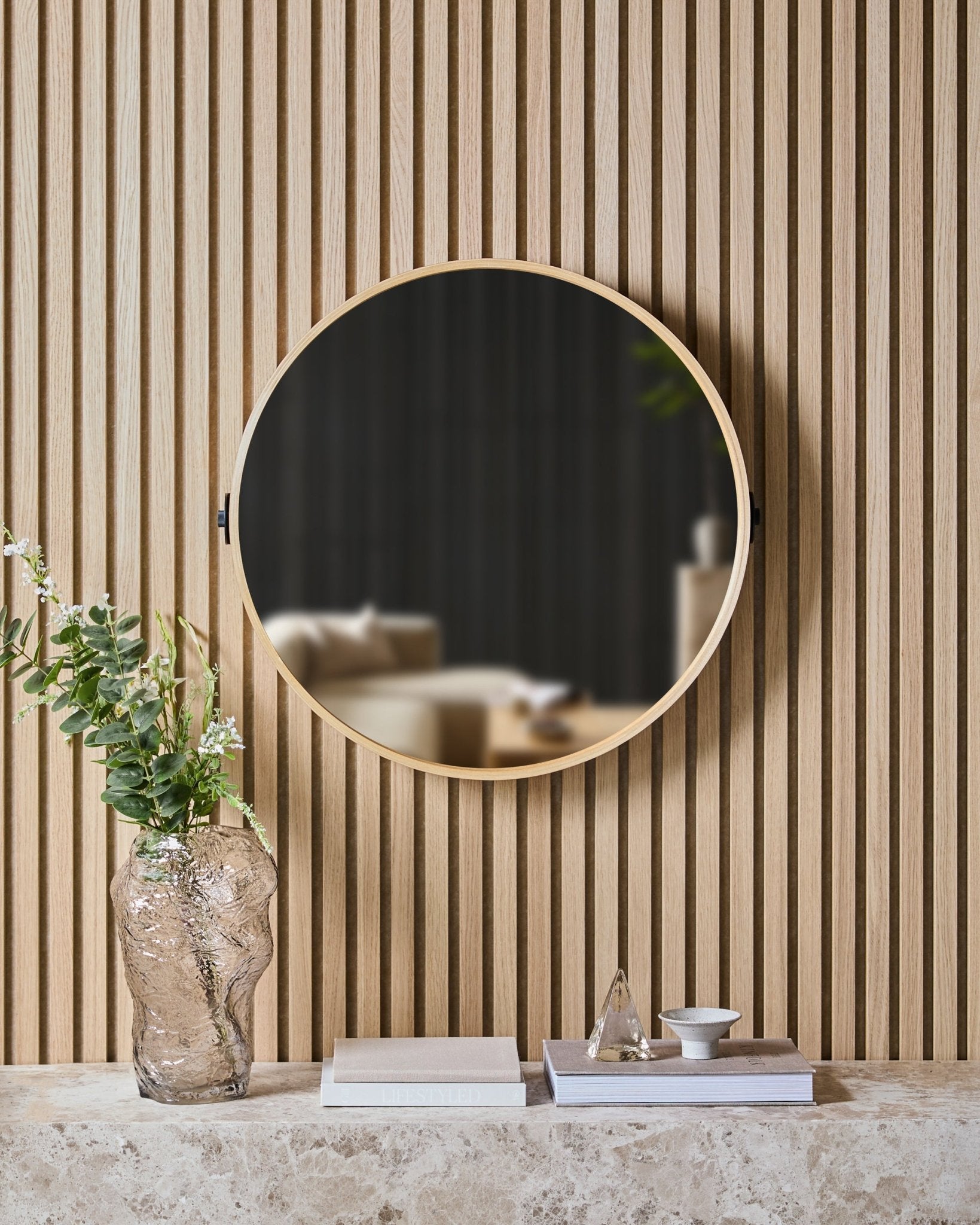 Der Linn | Mirror in Natural Oak von WoodUpp ist auf einem nordischen Lamellenwand-Akustikpaneel über einer Steinablage mit Glasvase, Büchern und Deko-Objekten montiert.