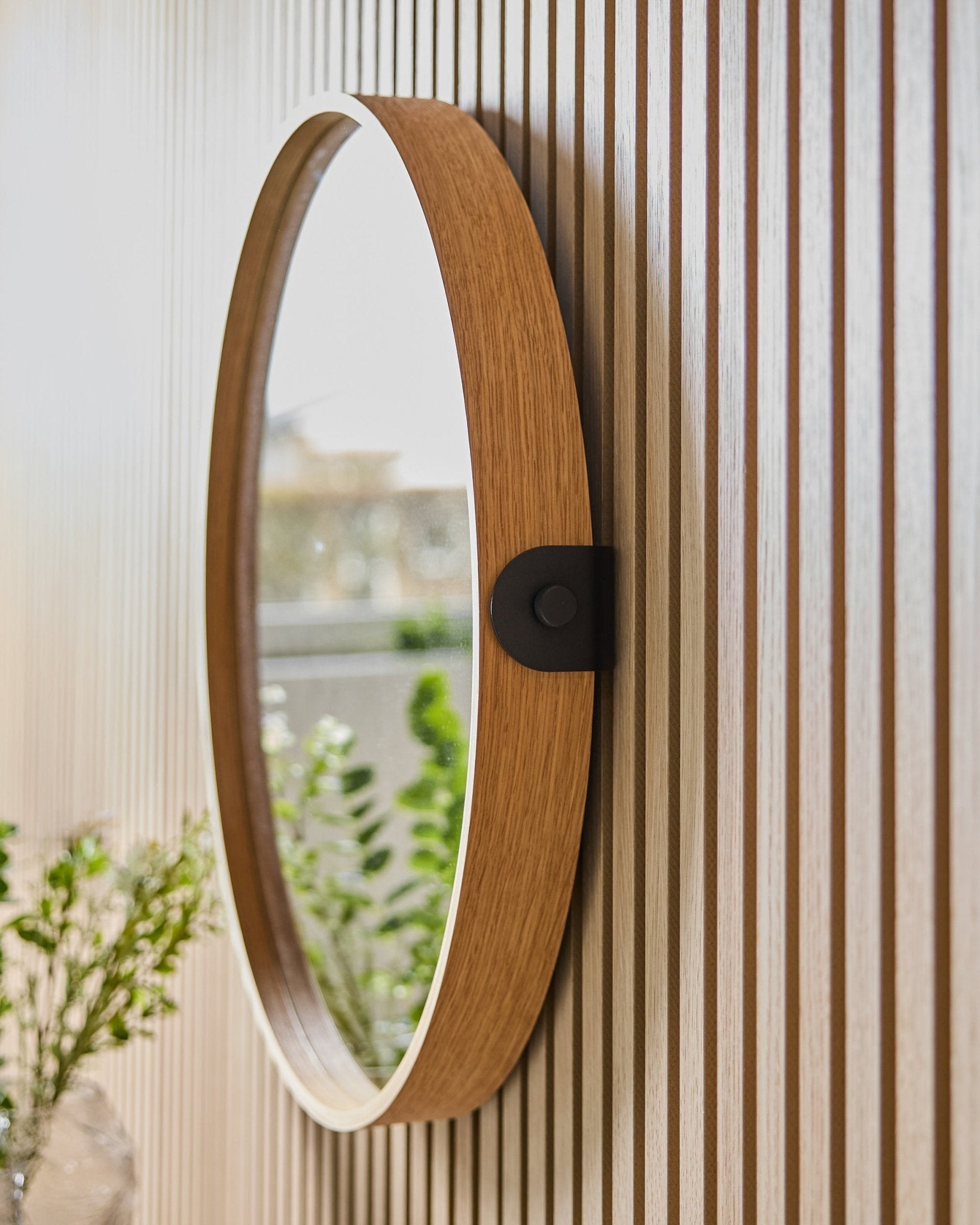 Linn | Mirror / Natural Oak von WoodUpp, montiert an einer gestreiften Akustikpaneel-Wand, die Grünpflanzen und natürliches Licht aus einem nahen Fenster reflektiert.