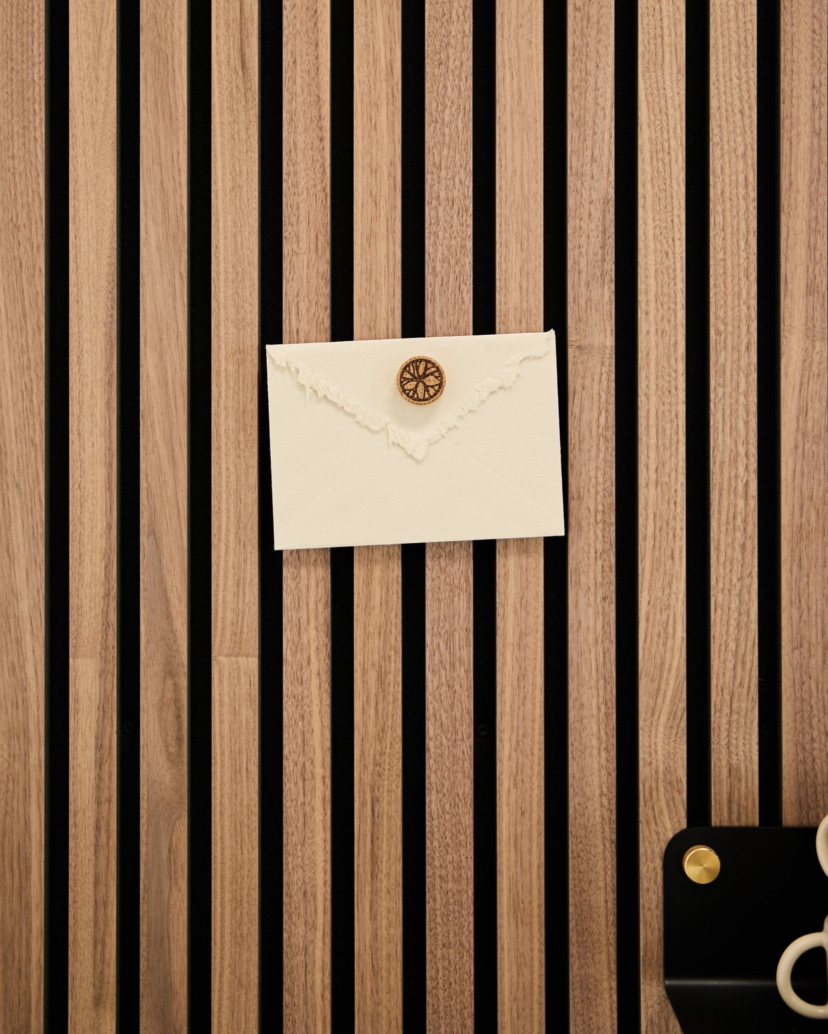 Ein cremefarbener Umschlag mit Wachssiegel ist mit dem Norr Poster Magnet Set in Natural Oak an einem WoodUpp Eichen-Akustik-Slatwall-Paneel befestigt; Schere und Tablett liegen daneben.