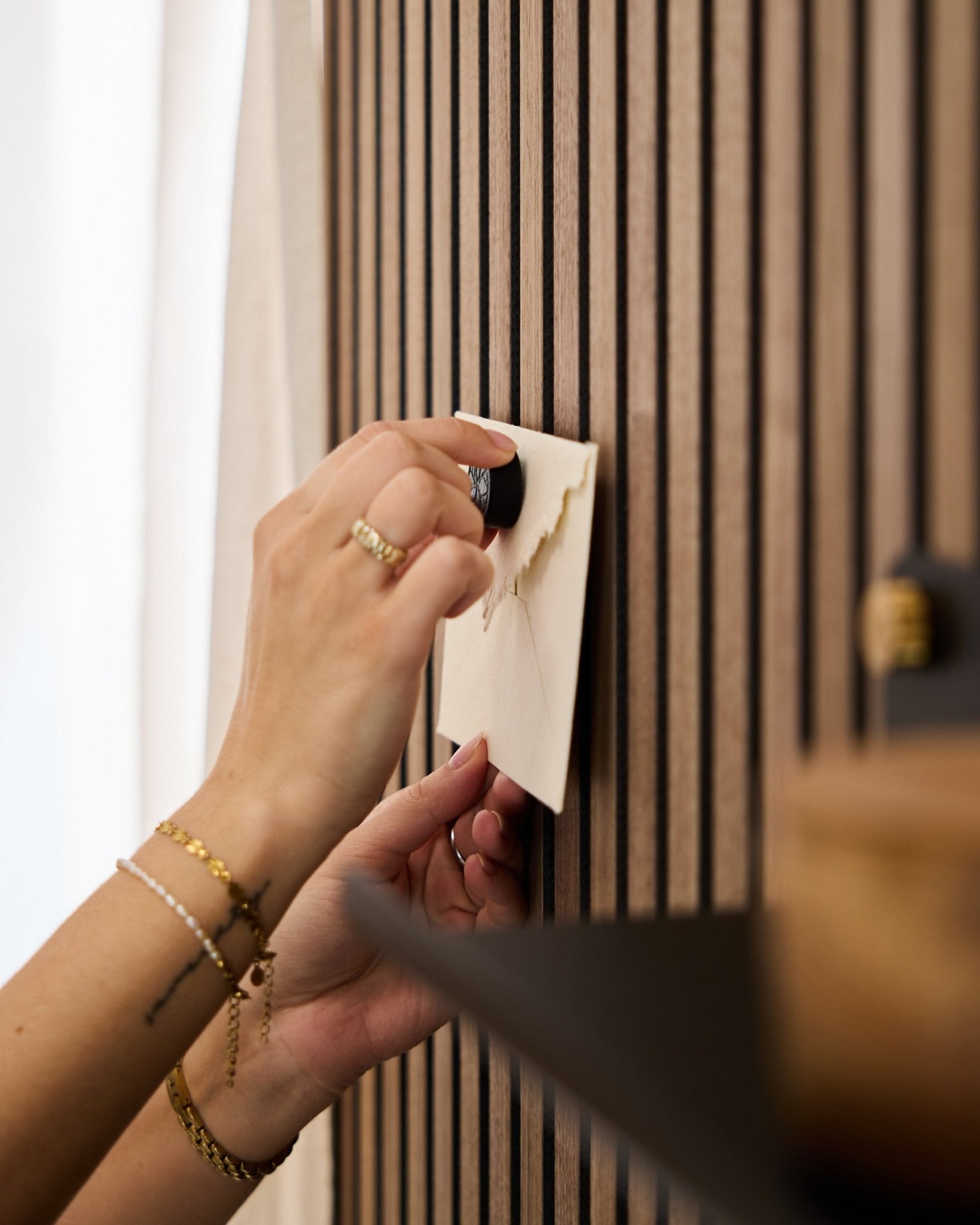 Eine Person mit goldenen Armbändern und Ringen befestigt mit dem Norr Poster-Magnet-Set in Black Oak einen Umschlag an einem WoodUpp Akustikpaneel mit Eichenlamellen.