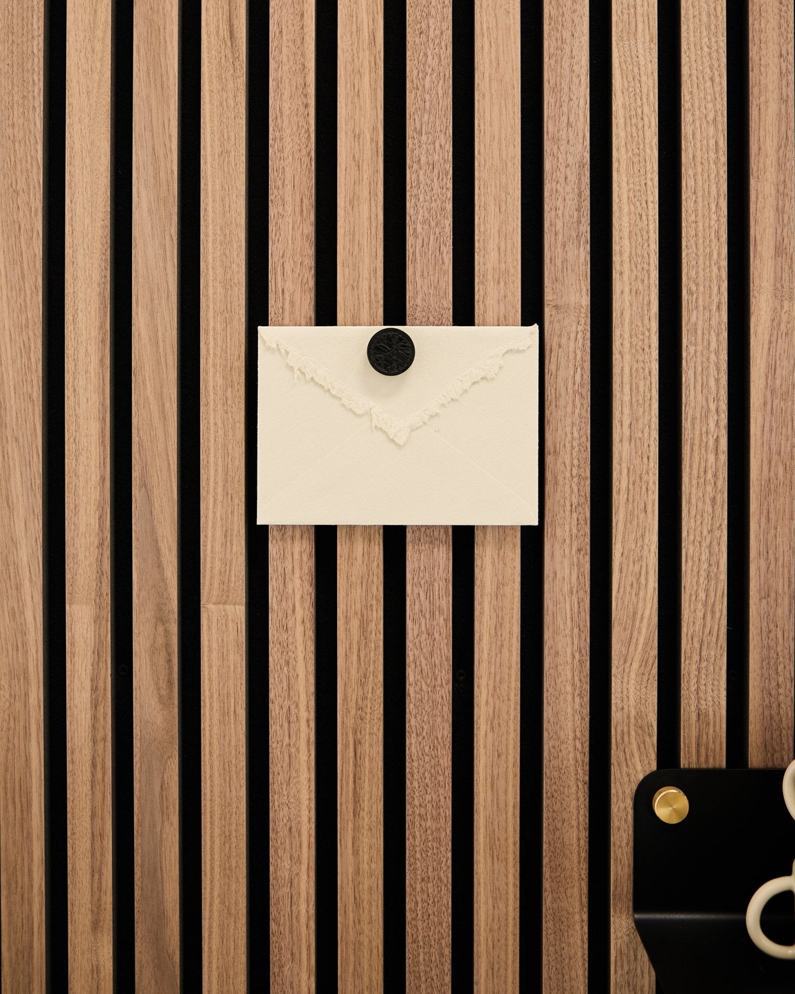 Une enveloppe crème est fixée à un Panneau acoustique WoodUpp à lattes en chêne Akupanel à l’aide du set d’aimants pour affiches Norr en Black Oak ; des ciseaux sont posés en bas à droite.