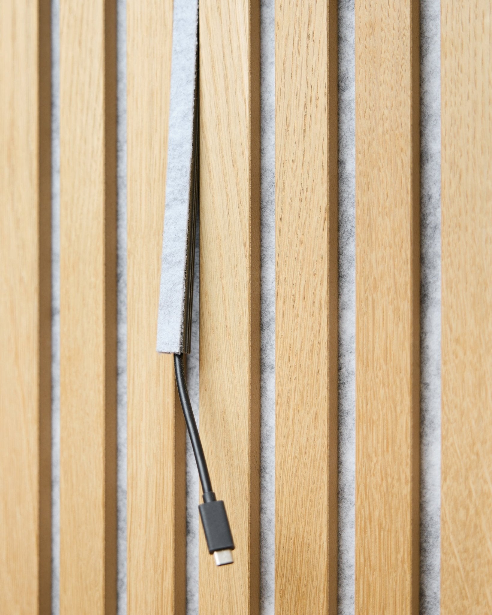 Ein black USB-C-Kabel verläuft durch eine grey WoodUpp Dupps Pop-on-Kabelabdeckung zwischen vertical Akustikholzlamellen mit grey Filzrückseite.