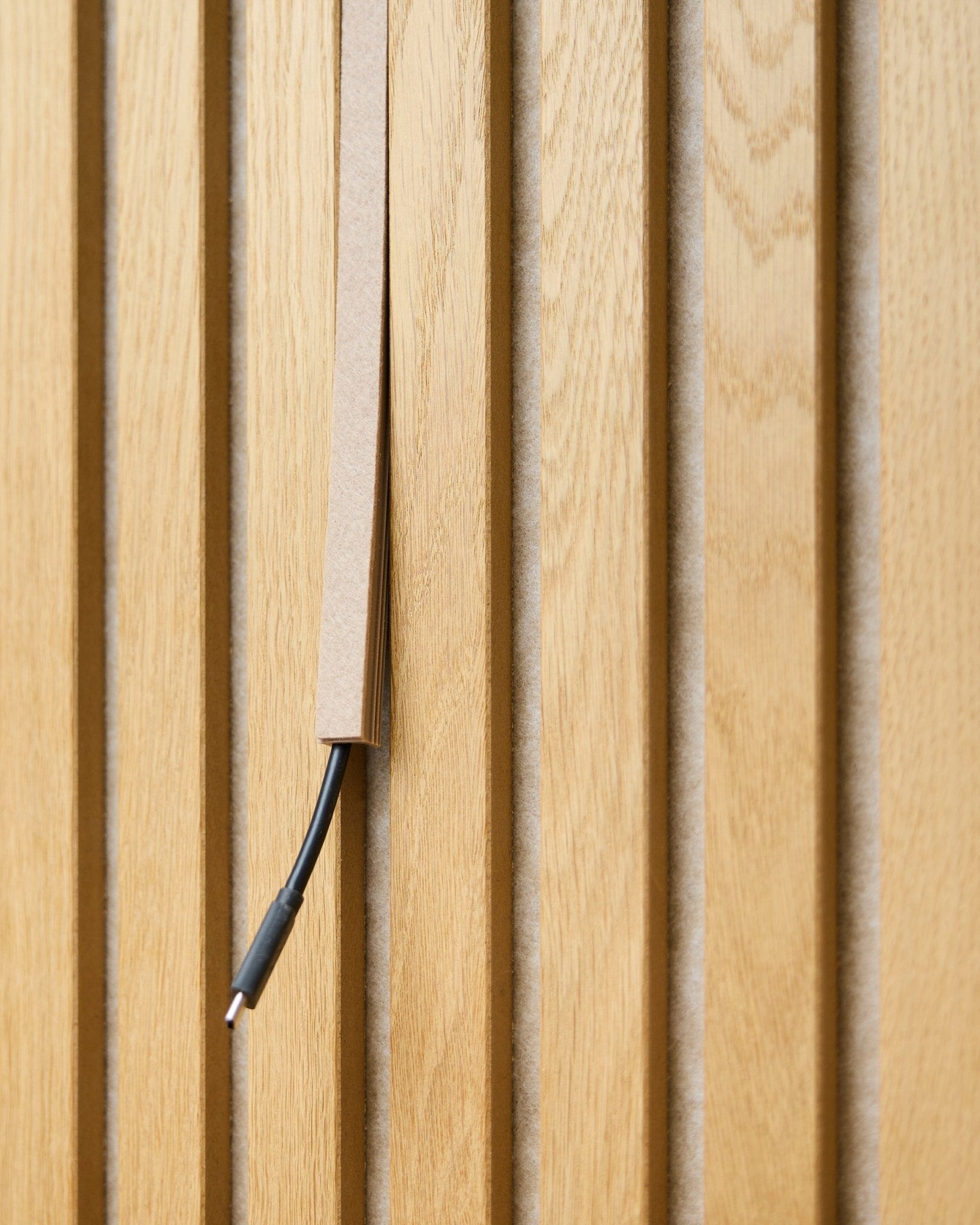 Un câble black avec gestion des câbles est partiellement dissimulé dans un WoodUpp Dupps Beige Pop-on Cable Cover, monté sur des lattes acoustiques en bois vertical Akupanel.
