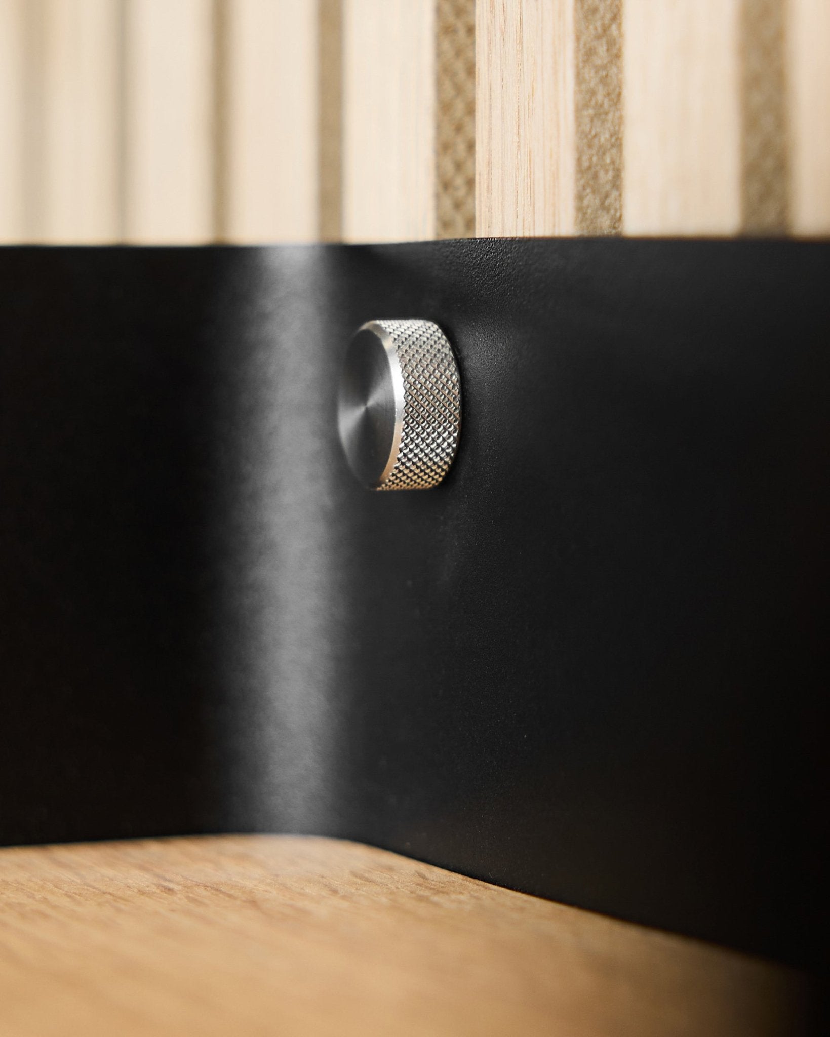 Nahaufnahme von zwei WoodUpp-Fingerschrauben (M5x10 mm, 20x8.8 aus rostfreiem steel) auf einer blacken Metalloberfläche, mit Akupanel Akustikholzlamellen im Hintergrund.