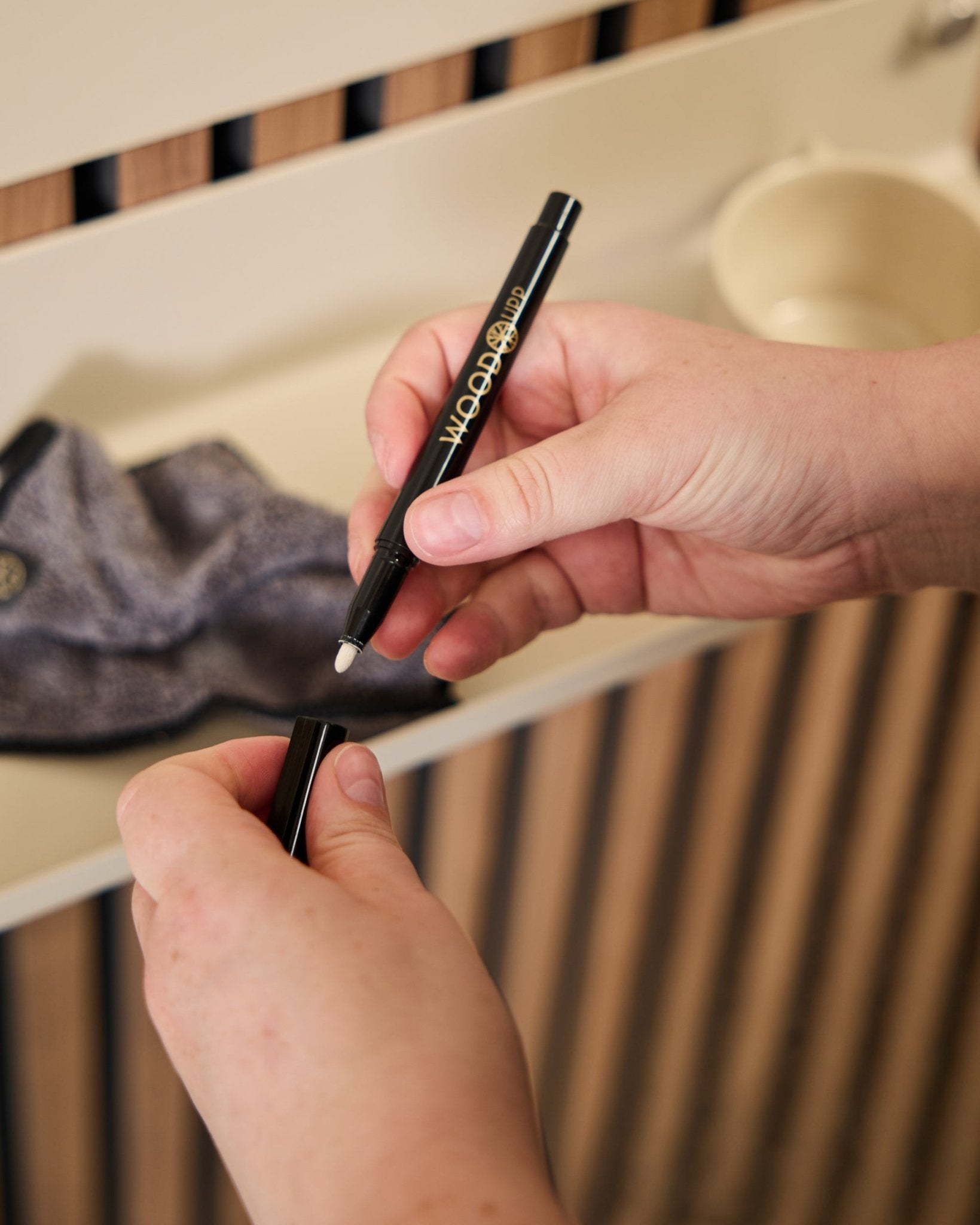 Eine Person hält einen geöffneten Marker des WoodUpp Norr | Blackboard set neben ein Mikrofasertuch und eine beige Tasse – Produkte, die für die Verwendung mit Akustik-Holzpaneelen entwickelt wurden.