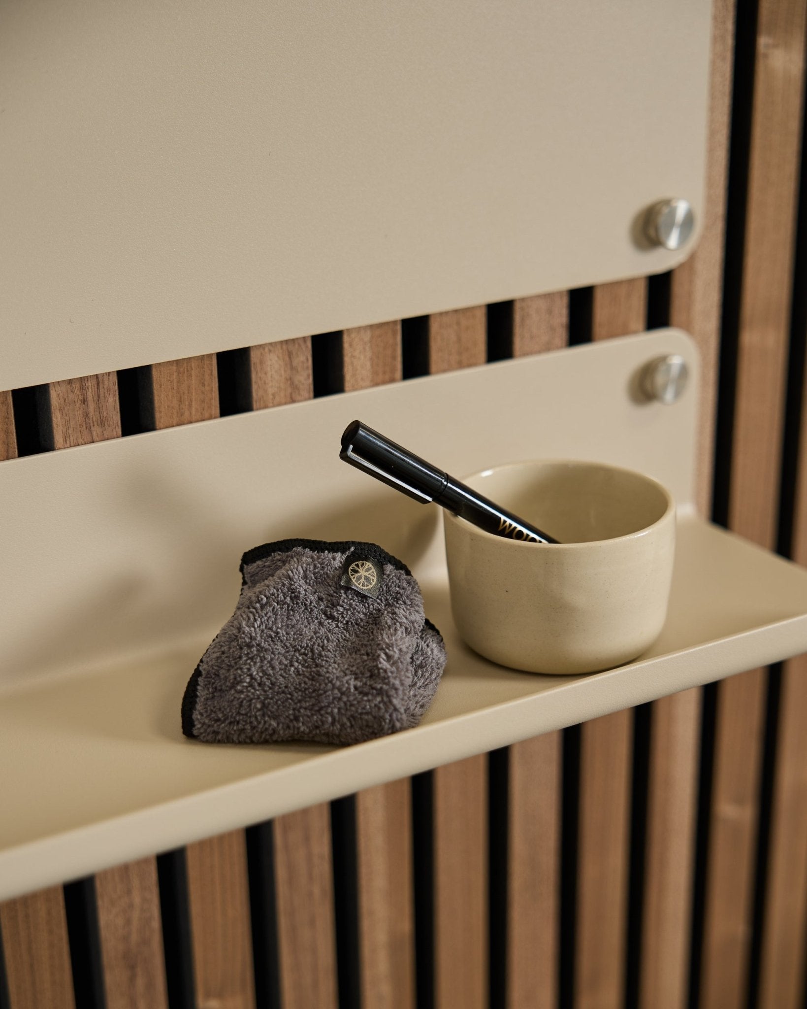 Das WoodUpp Norr Blackboard-Set umfasst eine smalle beige Tasse, eine Bürste mit blackem Griff und ein Reinigungstuch auf einem beigen Regal vor nordischer Akustik-Holzverkleidung.
