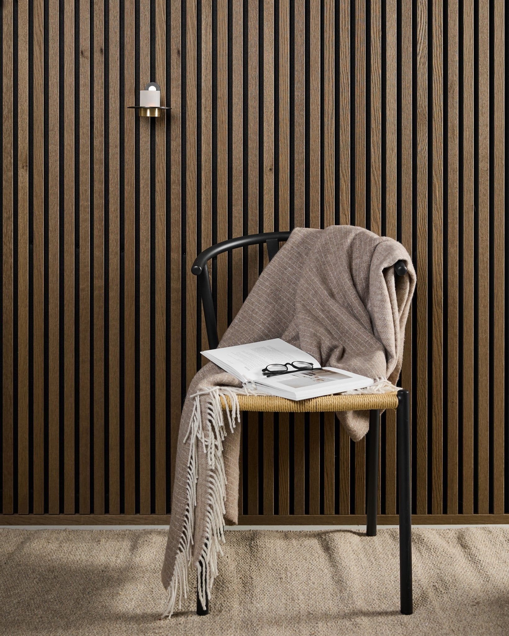 Ein moderner Stuhl mit einer beigefarbenen Wolldecke, einem aufgeschlagenen Buch und einer Brille steht neben einem Akupanel Akustikholzpaneel und einem WoodUpp black- und brassfarbenen Norr Kerzenhalter.