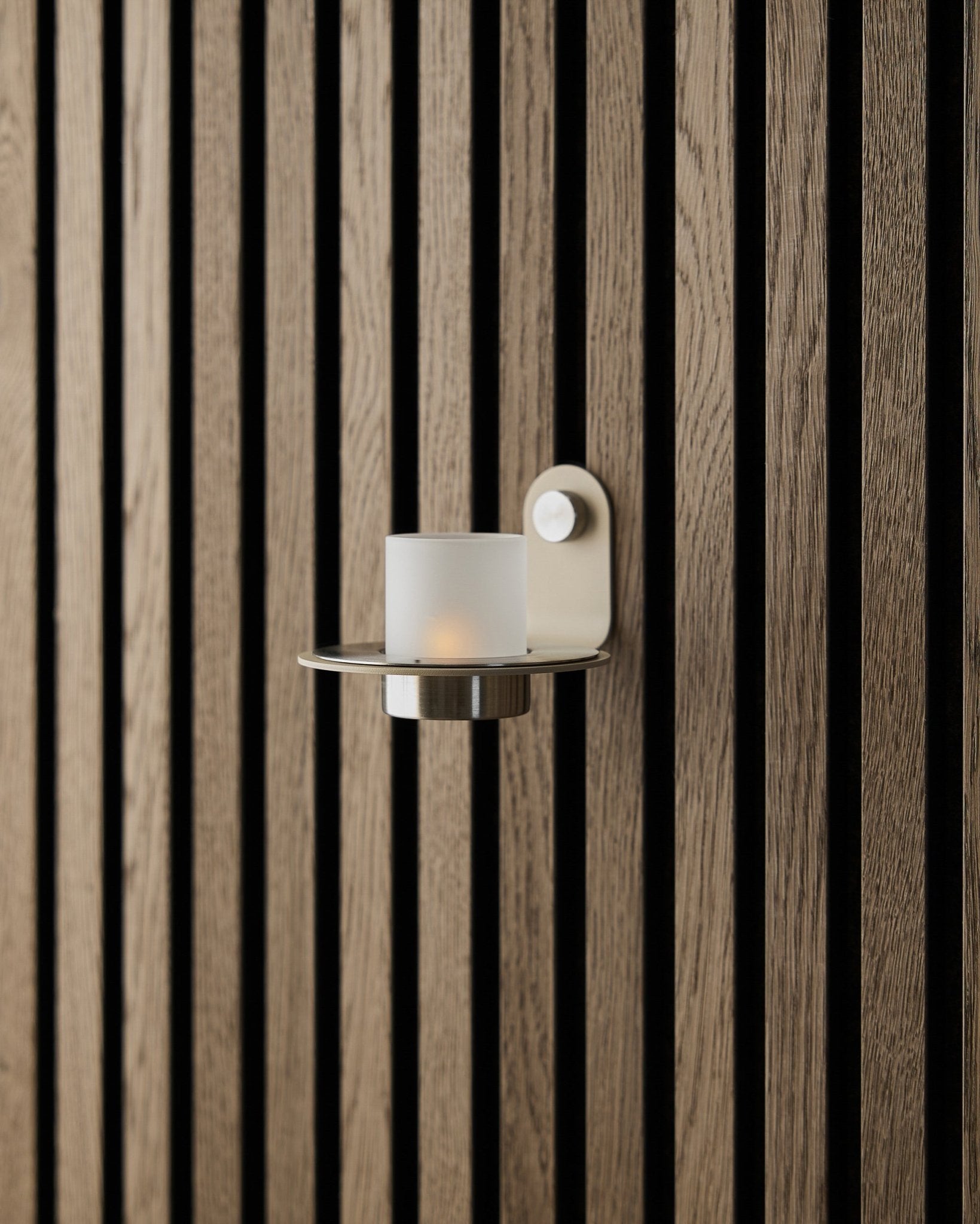 Der Norr-Kerzenhalter aus beigefarbenem Edelstahl von WoodUpp, präsentiert vor einer beigefarbenen Akupanel Akustikpaneel-Wand.