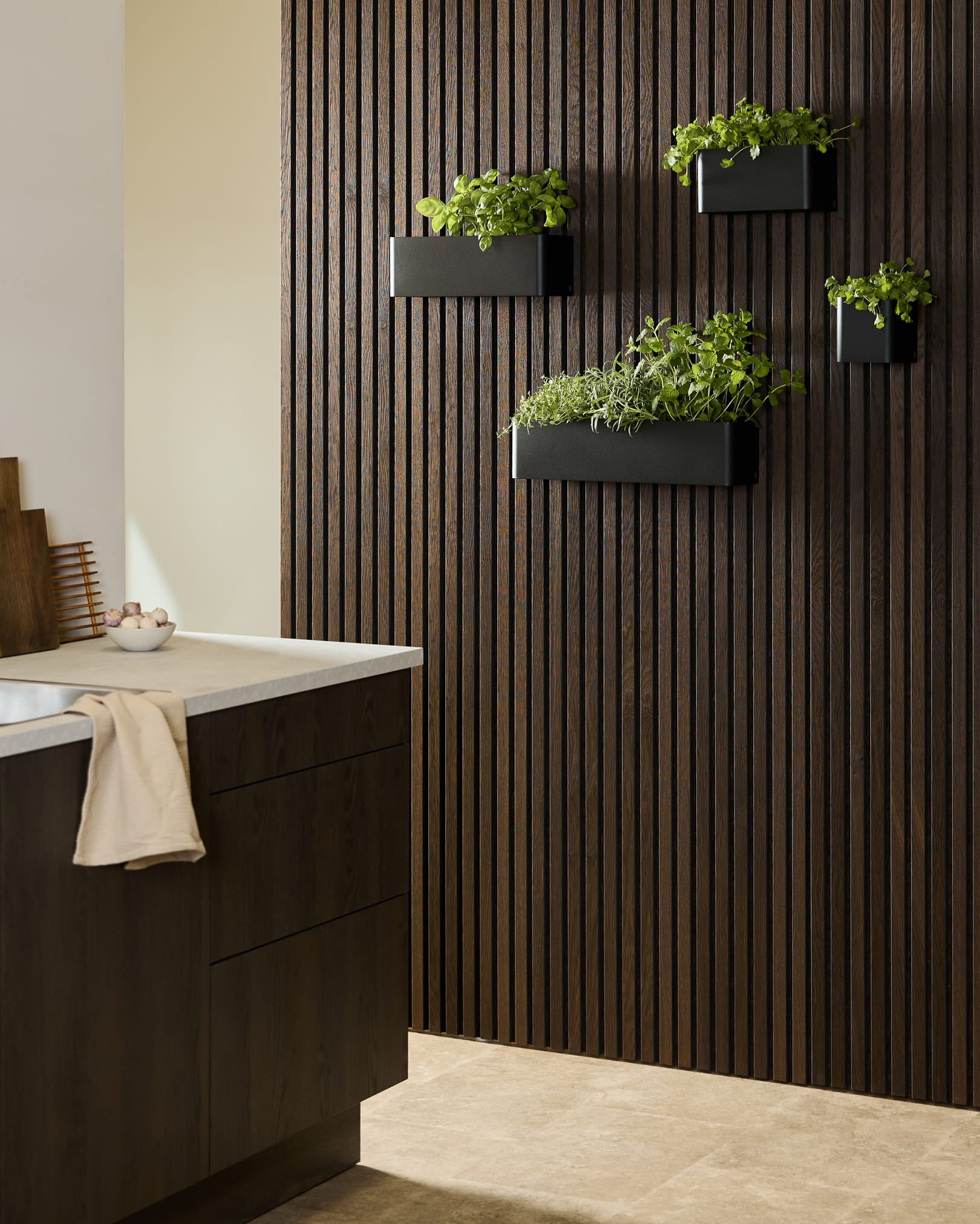Cuisine moderne avec un Akupanel en bois foncé de WoodUpp, un Panneau acoustique, un black Norr Plant Box avec des herbes, et des placards foncés surmontés d’un plan de travail blanc.