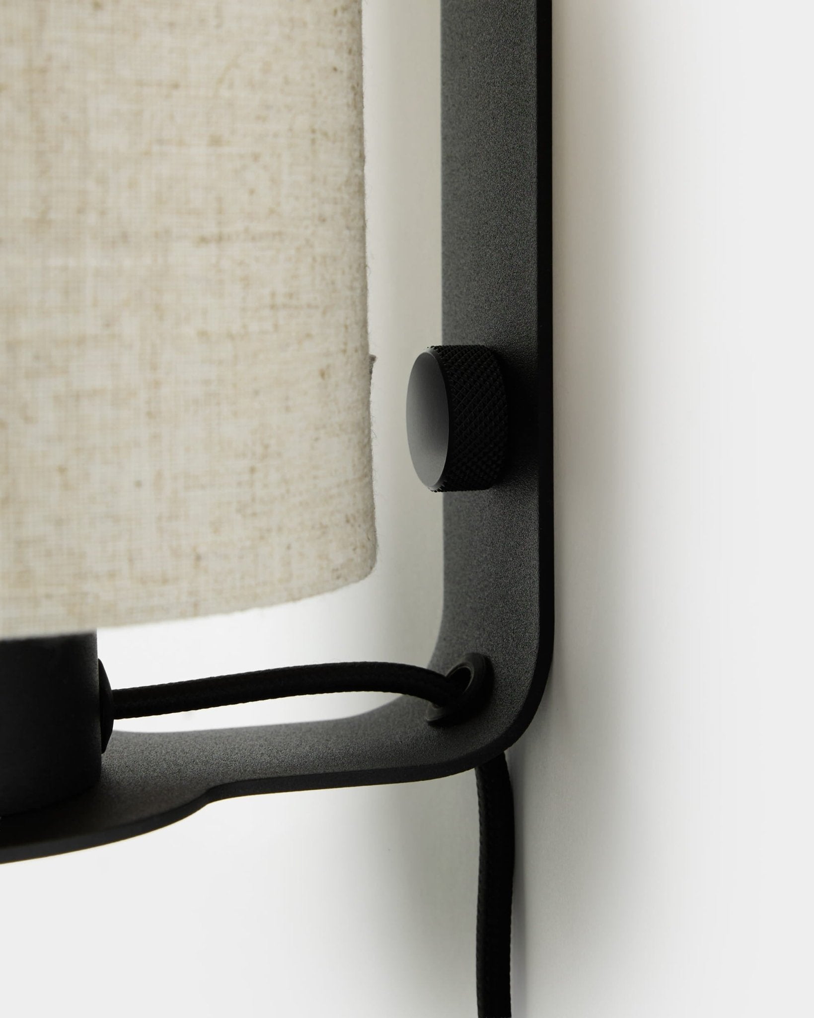 Gros plan sur l’applique murale Feya de WoodUpp en black et beige, avec bouton texturé, abat-jour en lin-coton et cordon visible, montée sur un mur blanc.